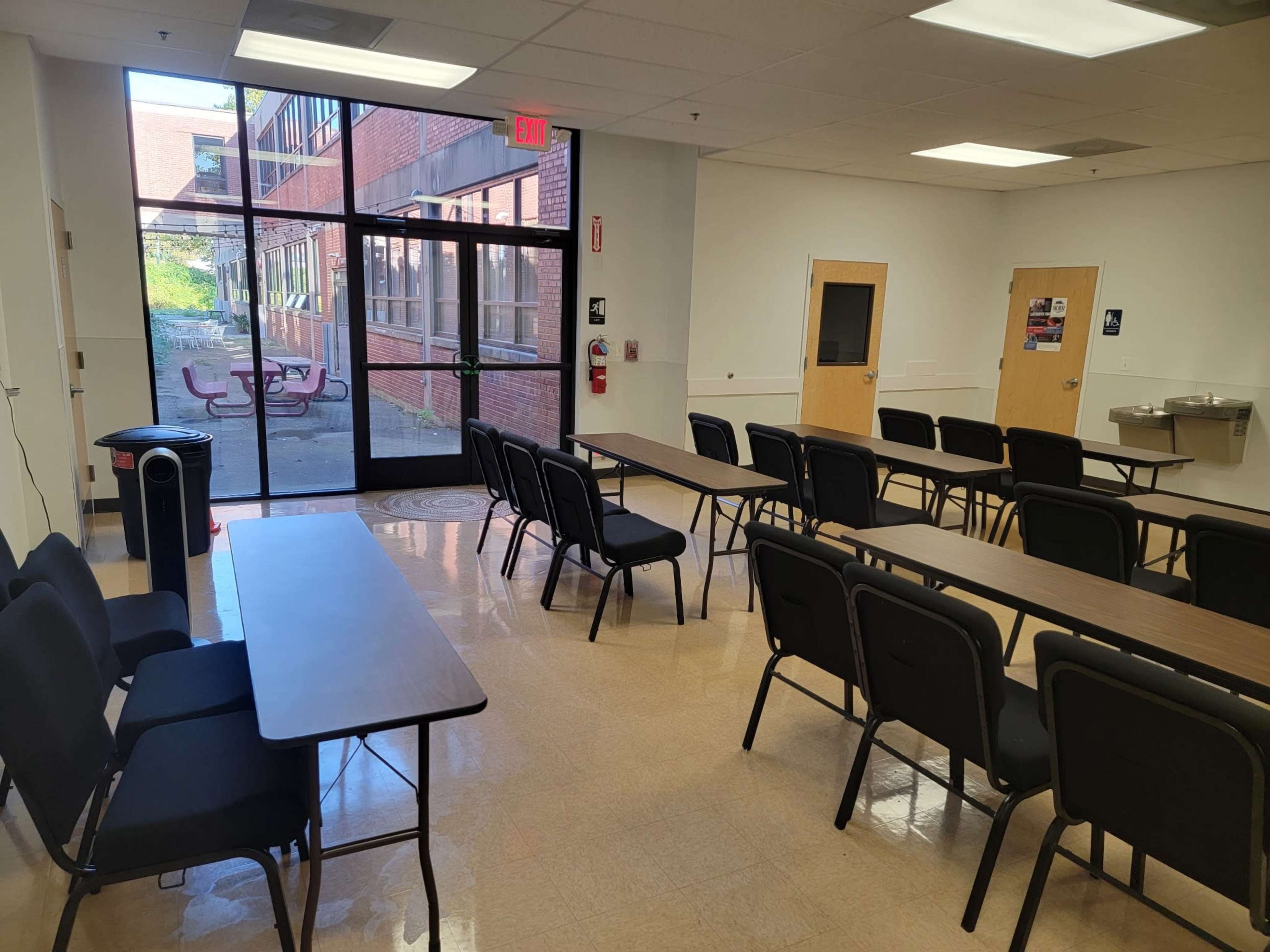 The image shows a large, brightly lit room with several rows of chairs and tables, along with a water fountain and glass doors leading to an outdoor area.
