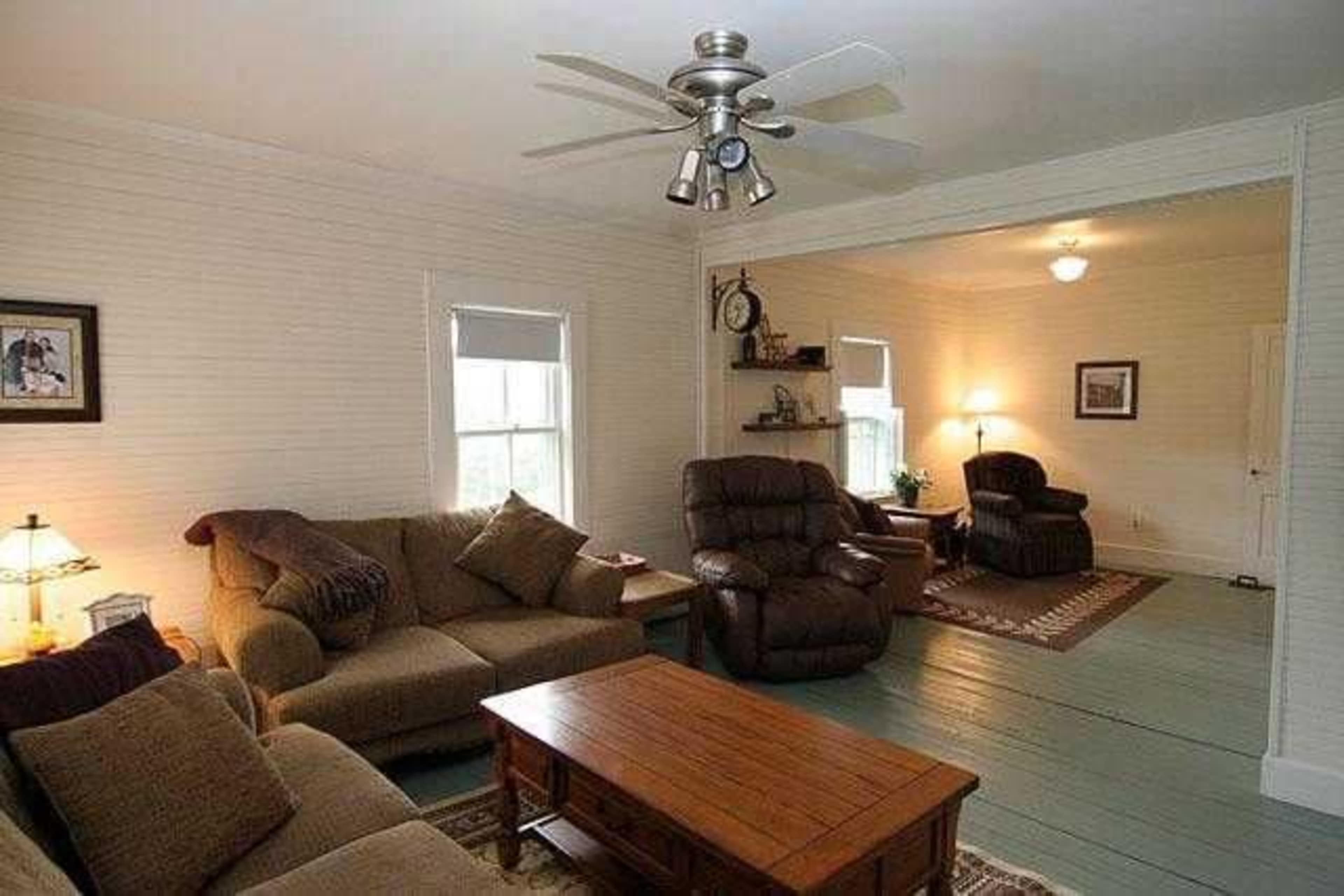 A living room with a couch, recliner chairs, a wooden coffee table, and light-colored walls, connected to another room through an archway.