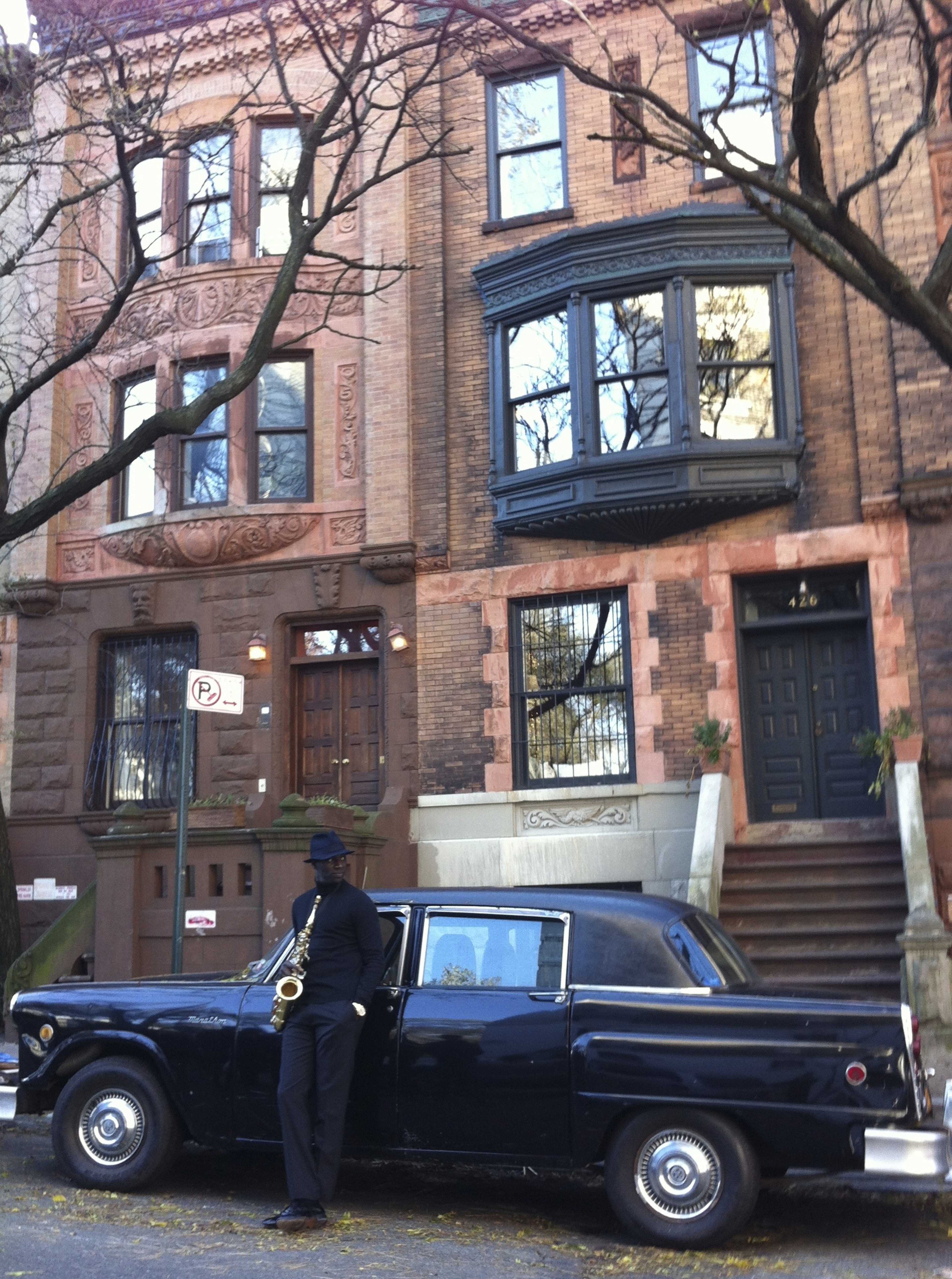 A man playing a saxophone stands next to an old black car in front of a brownstone building with intricate architectural details.