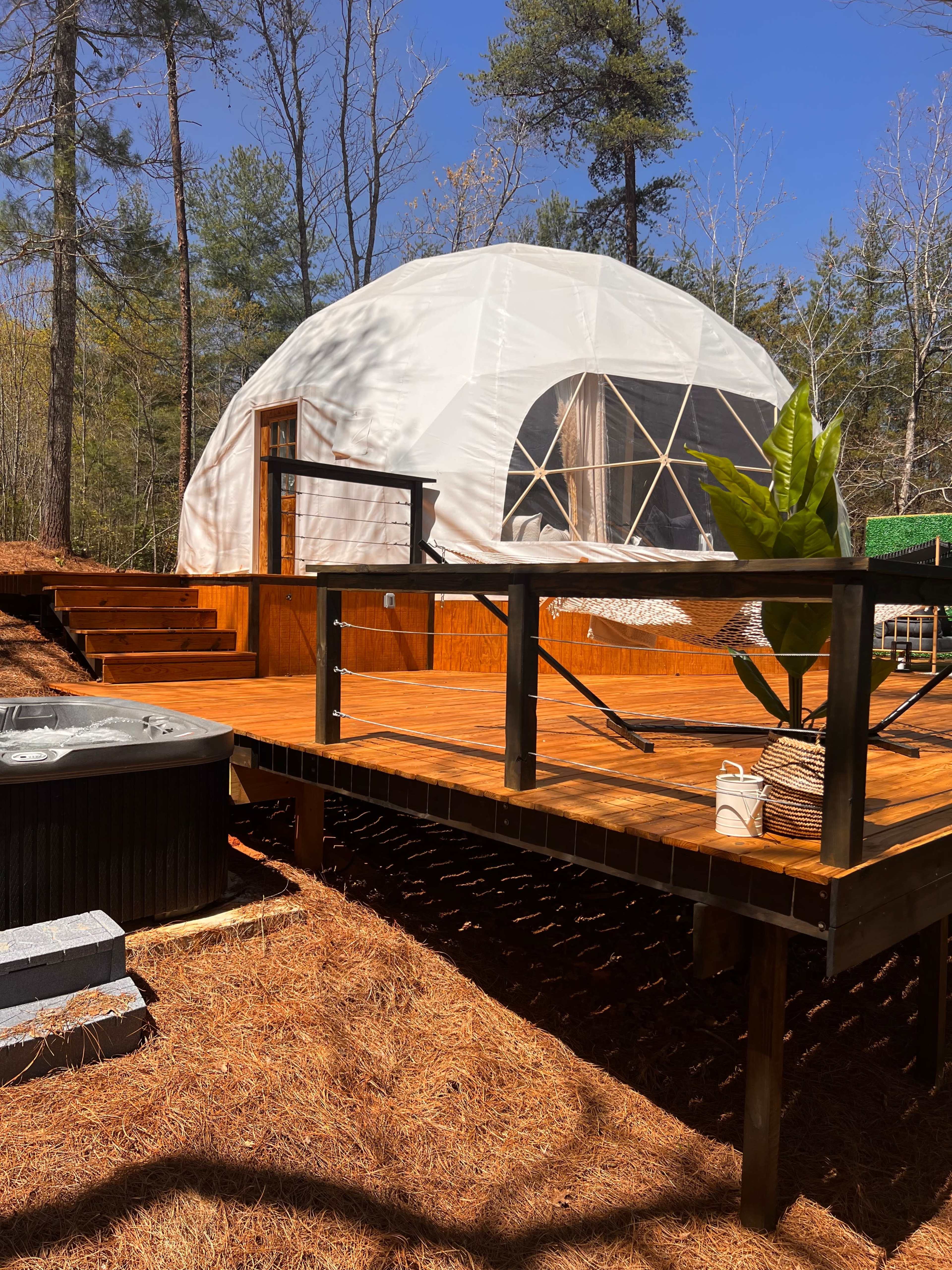 A geodesic dome is situated on a wooden deck surrounded by trees, with a hot tub nearby.