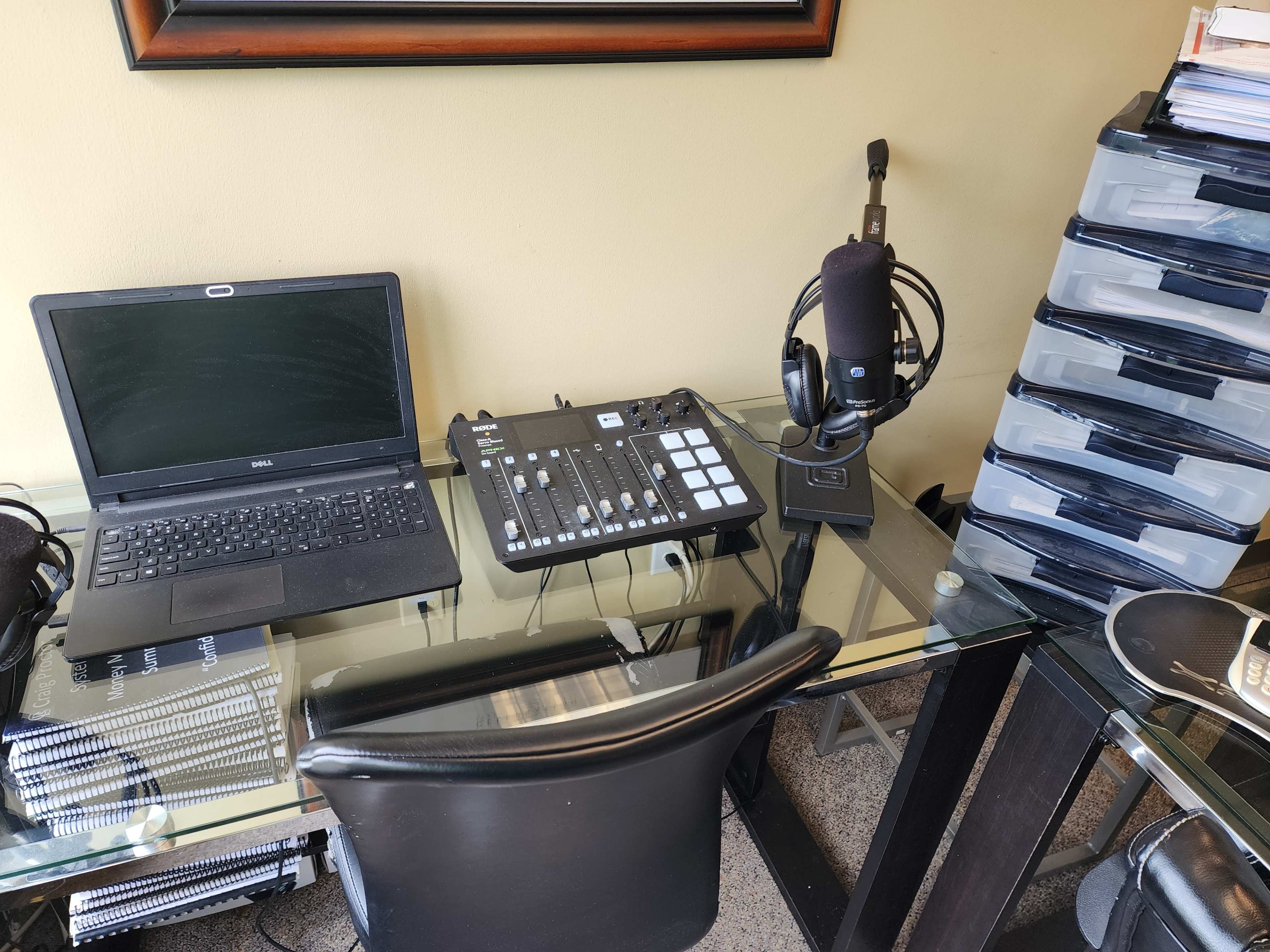 A home office setup features a laptop, audio mixer, microphone, and storage drawers on a glass desk.