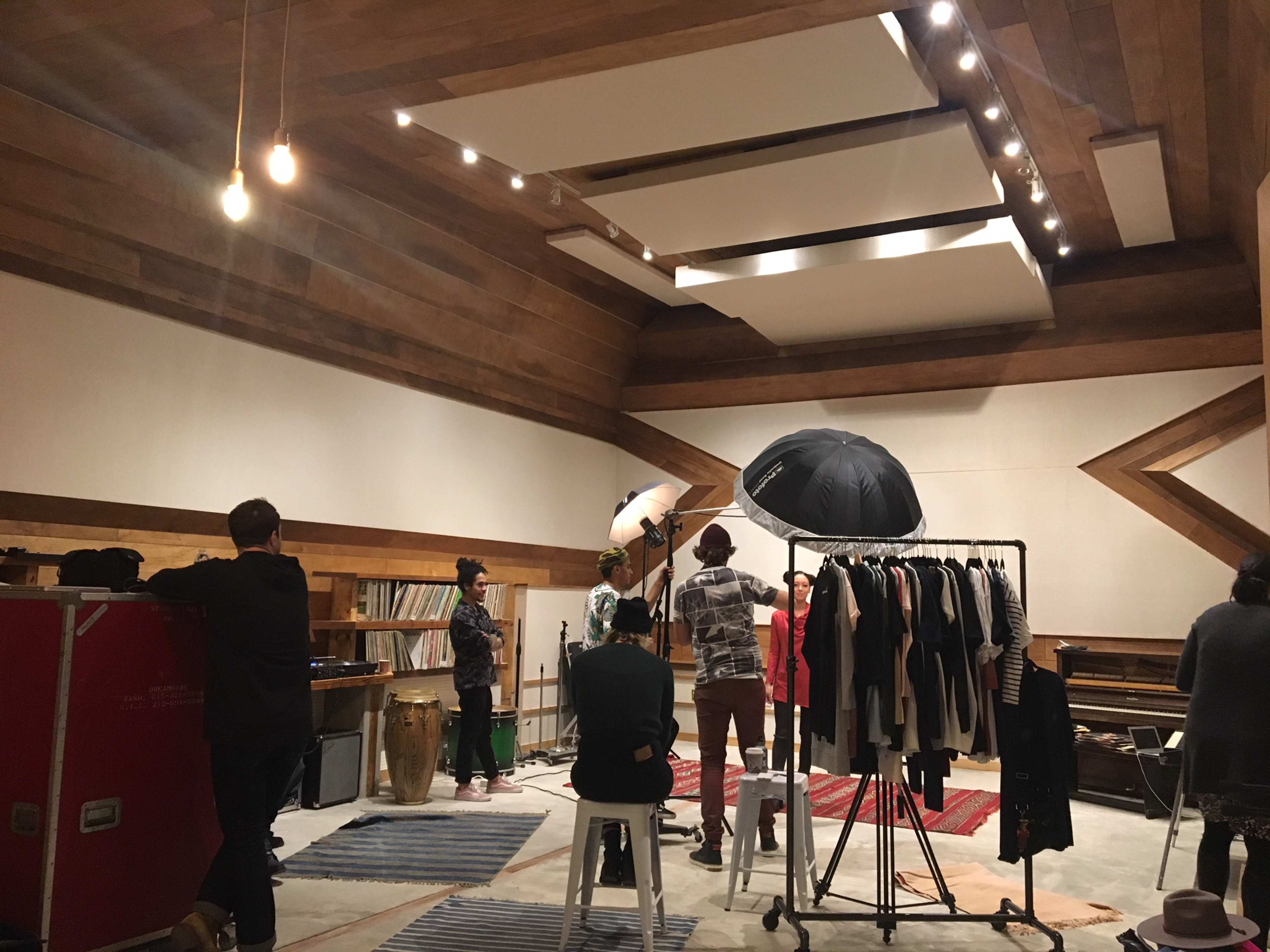 A group of people stands in a wooden studio space arranged for a photoshoot, with clothing on racks, musical instruments, and soft lighting fixtures.