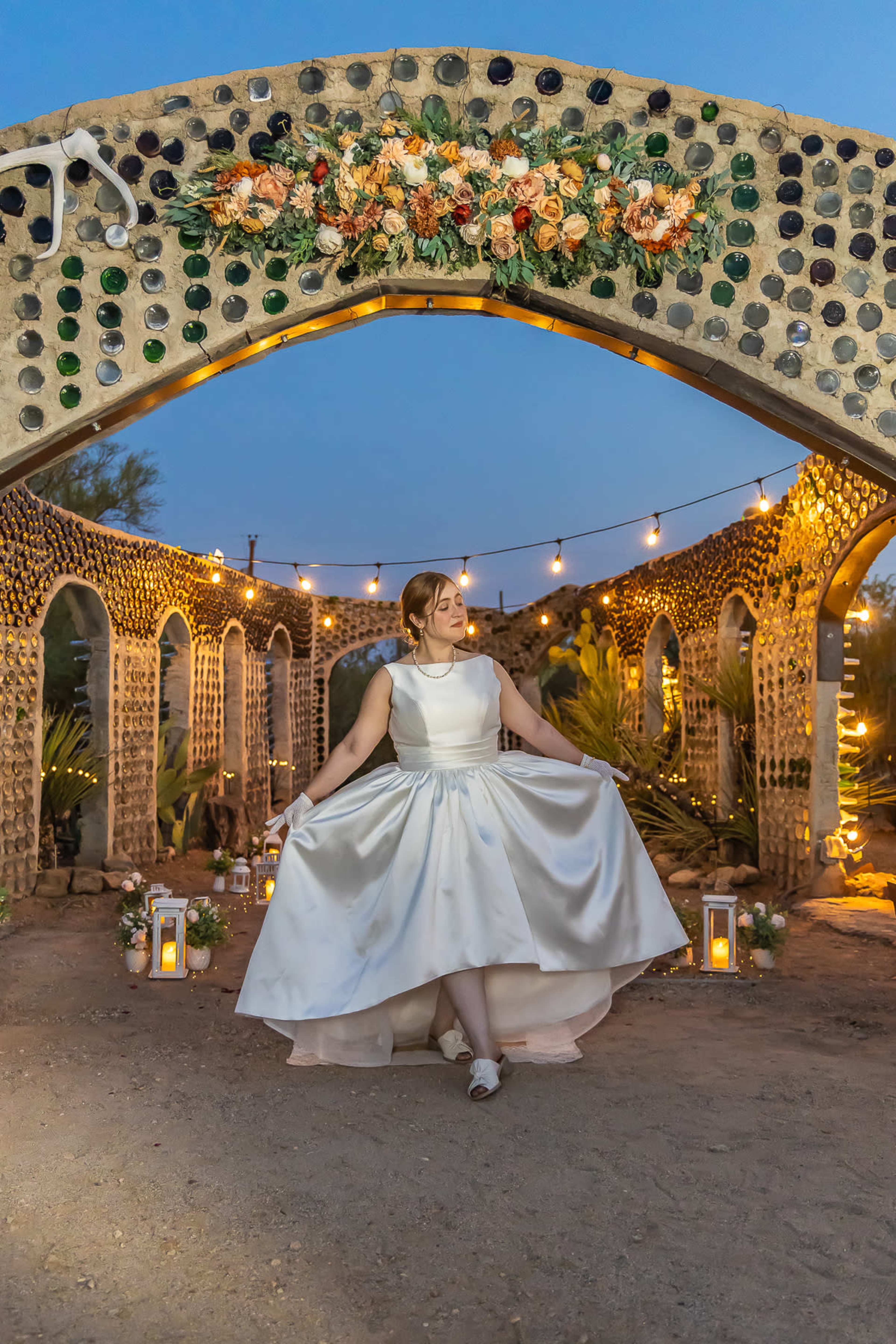 A bride in a white satin dress poses under an arch adorned with flowers and lights at an outdoor venue.