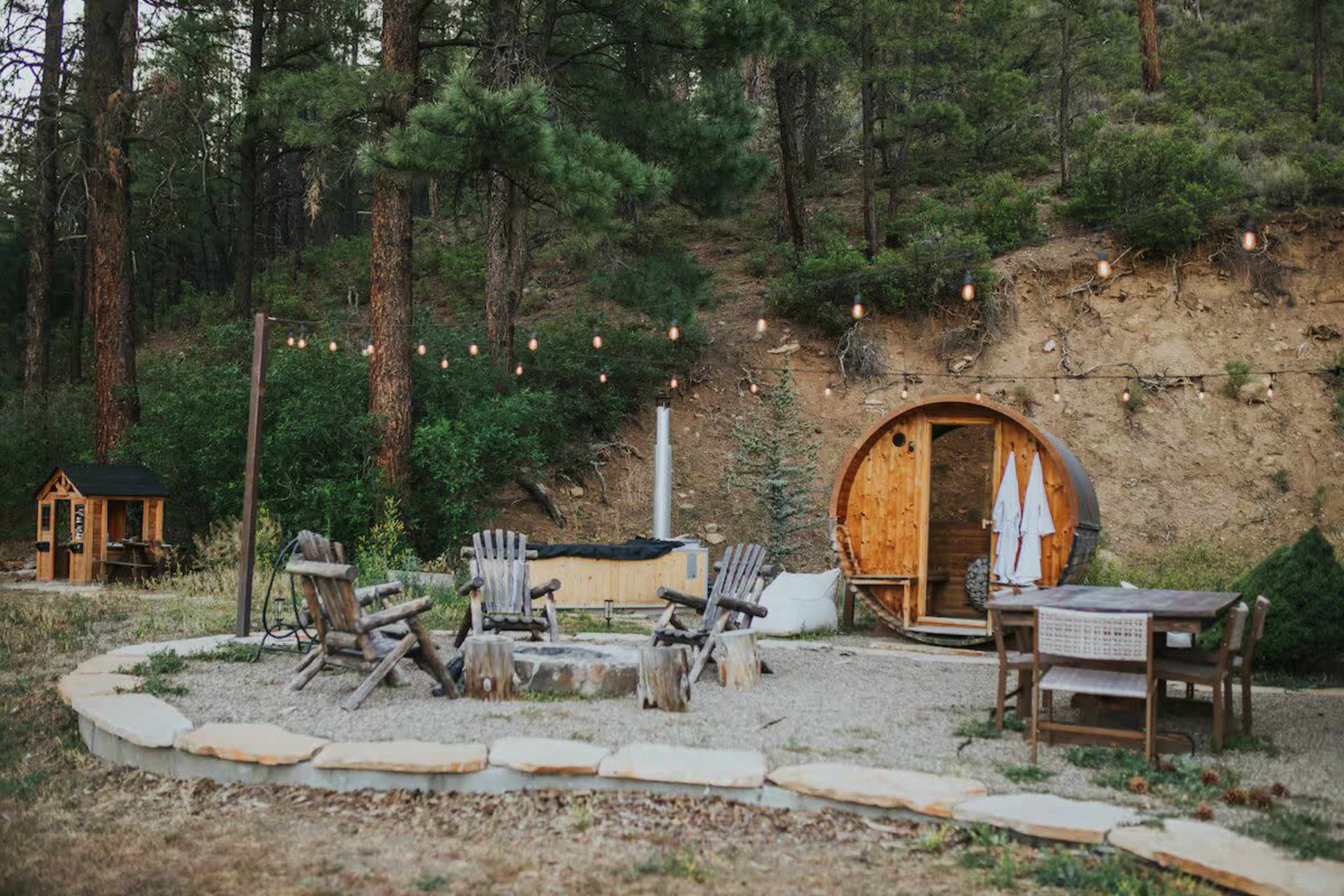 The image shows a rustic outdoor gathering area with a circular stone fire pit, wooden seating, and a barrel-shaped structure beside a forested background.