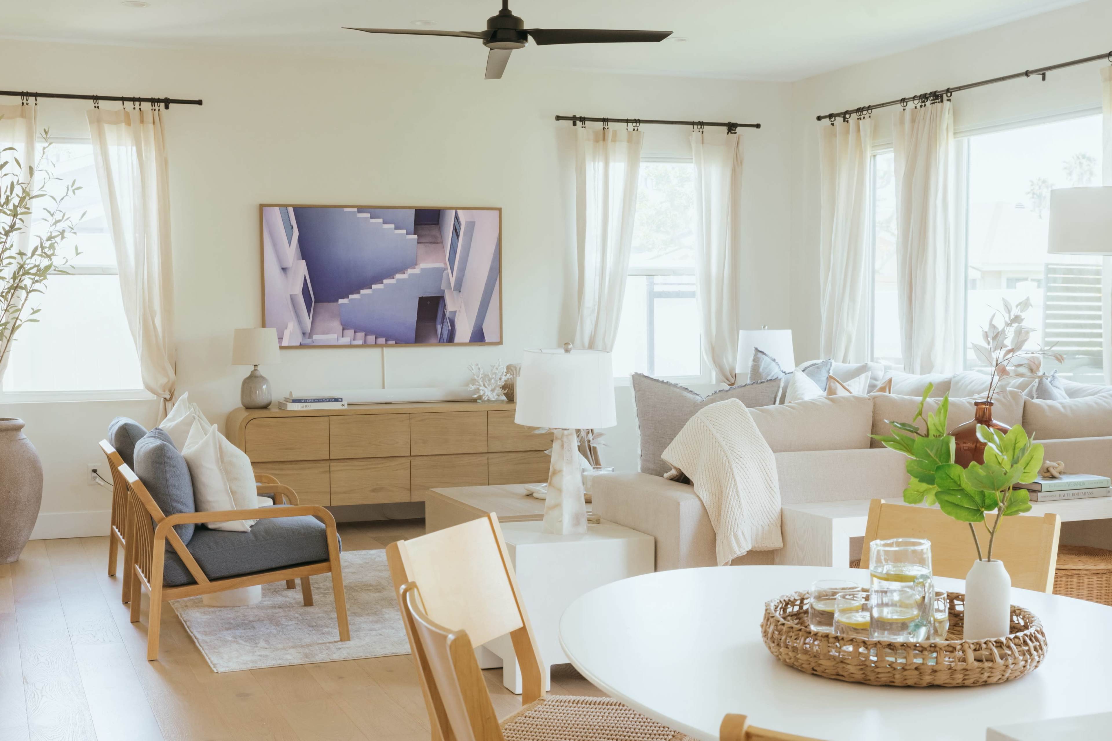 A modern living room features a neutral color palette, a large television mounted on the wall, and a combination of seating areas with natural light streaming through the windows.