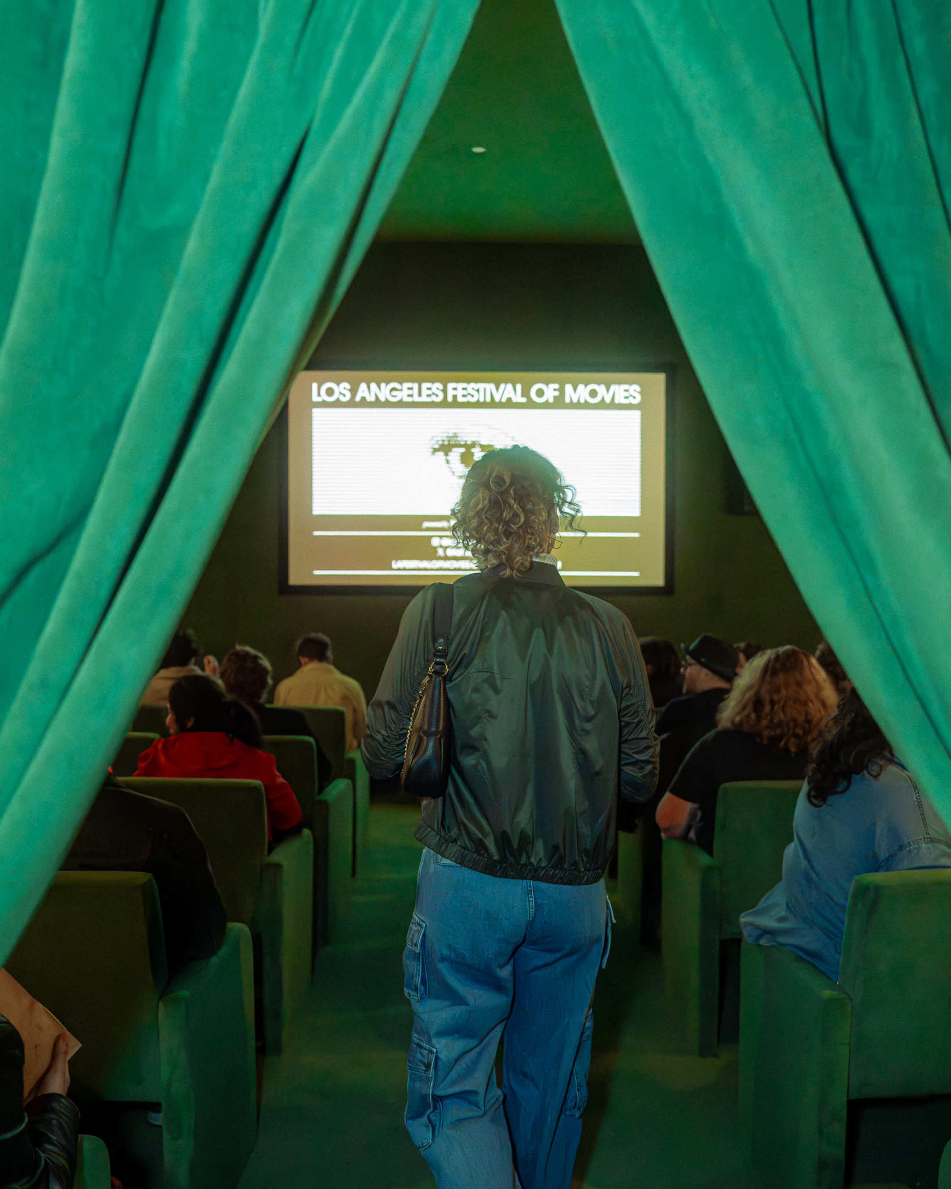 A person stands at the entrance of a theater, viewing a screen that displays "Los Angeles Festival of Movies" while an audience is seated in green chairs.