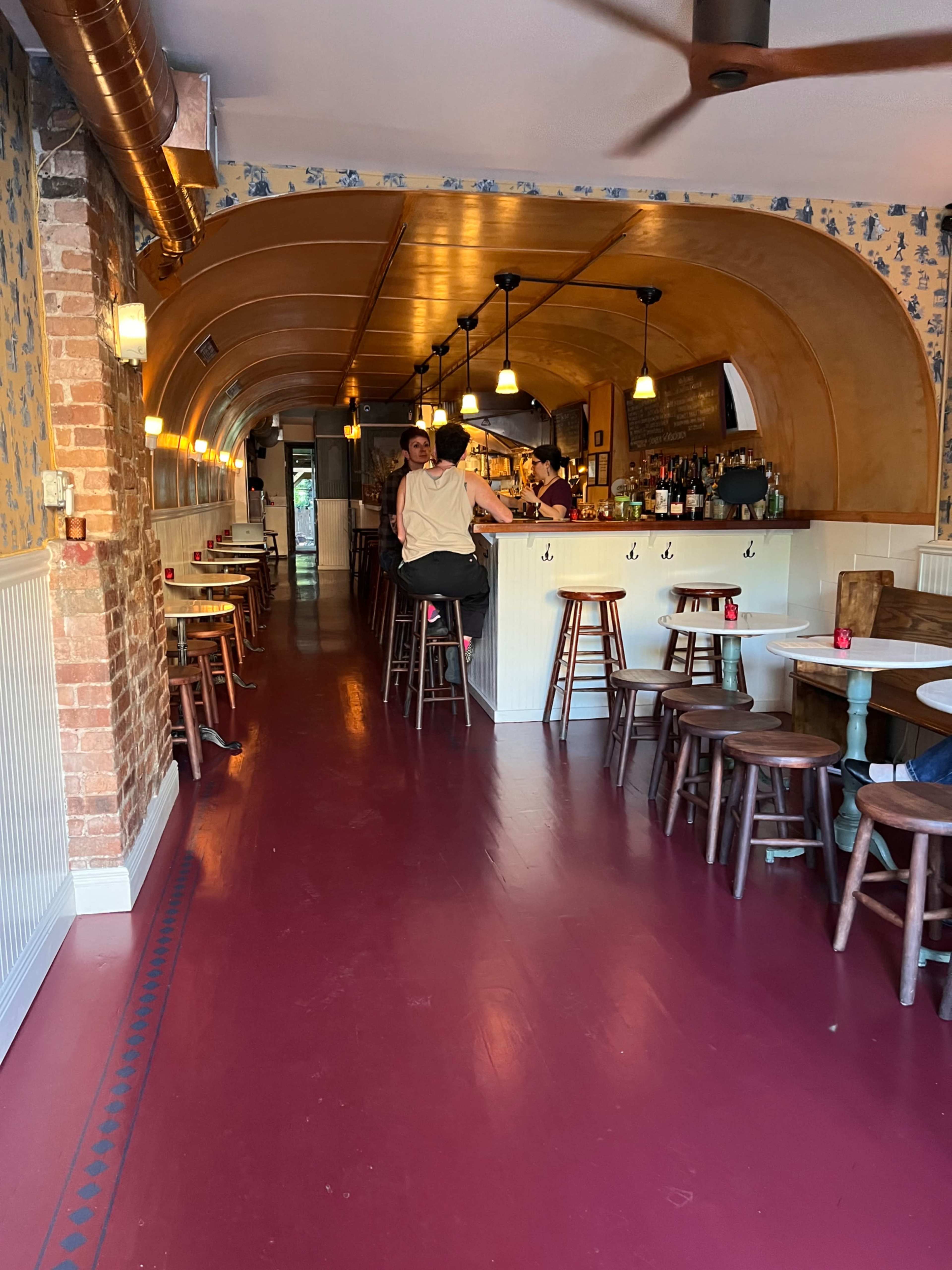 The image shows a narrow restaurant interior with wooden tables, bar stools, and a curved ceiling, leading to a bar area in the back.