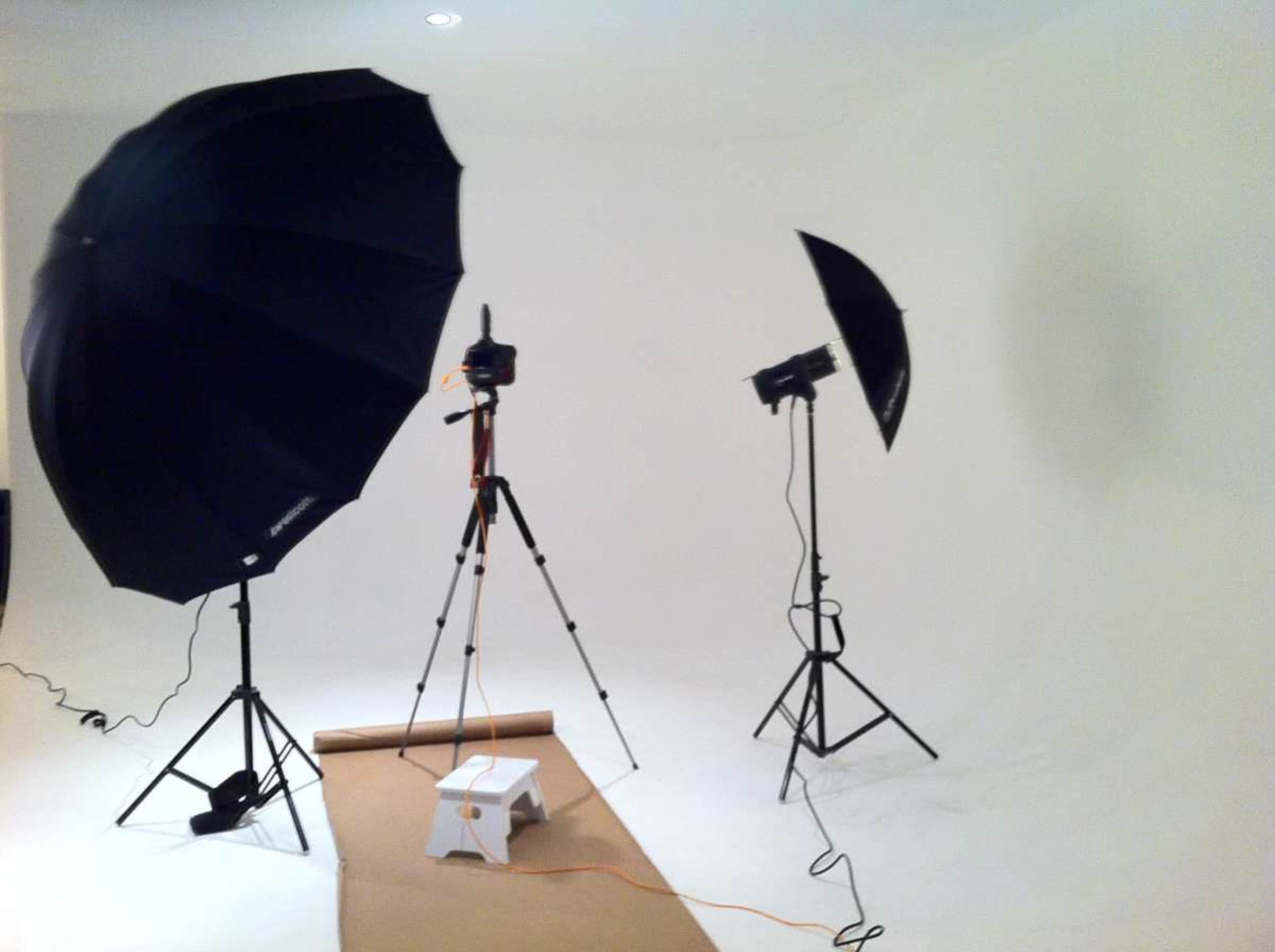 The image shows a photography studio setup with two light umbrellas, a camera on a tripod, and a small step stool on a brown paper backdrop against a plain white wall.