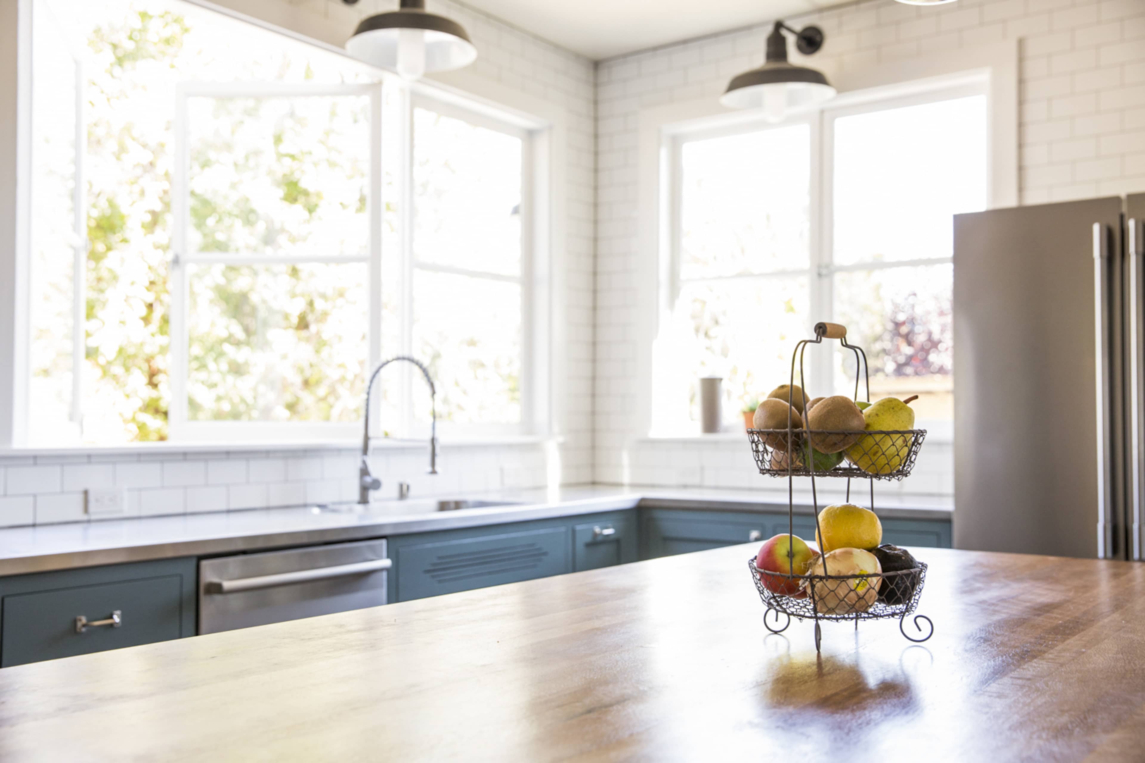 A modern kitchen features a wooden counter with a wire fruit basket filled with various fruits, large windows allowing natural light to brighten the space.