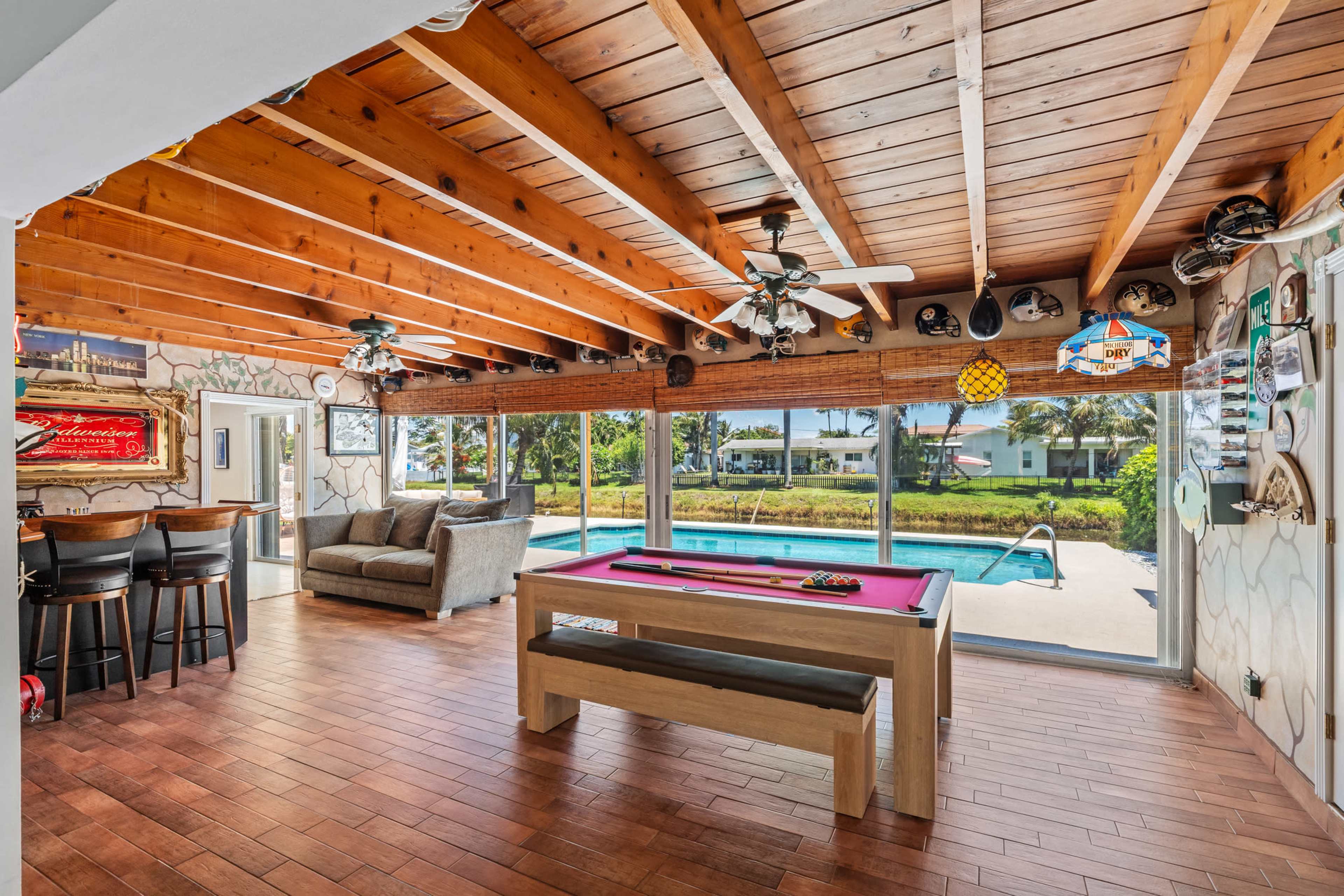 The image shows an indoor space with a pool table, seating area, and large windows overlooking a pool and backyard.
