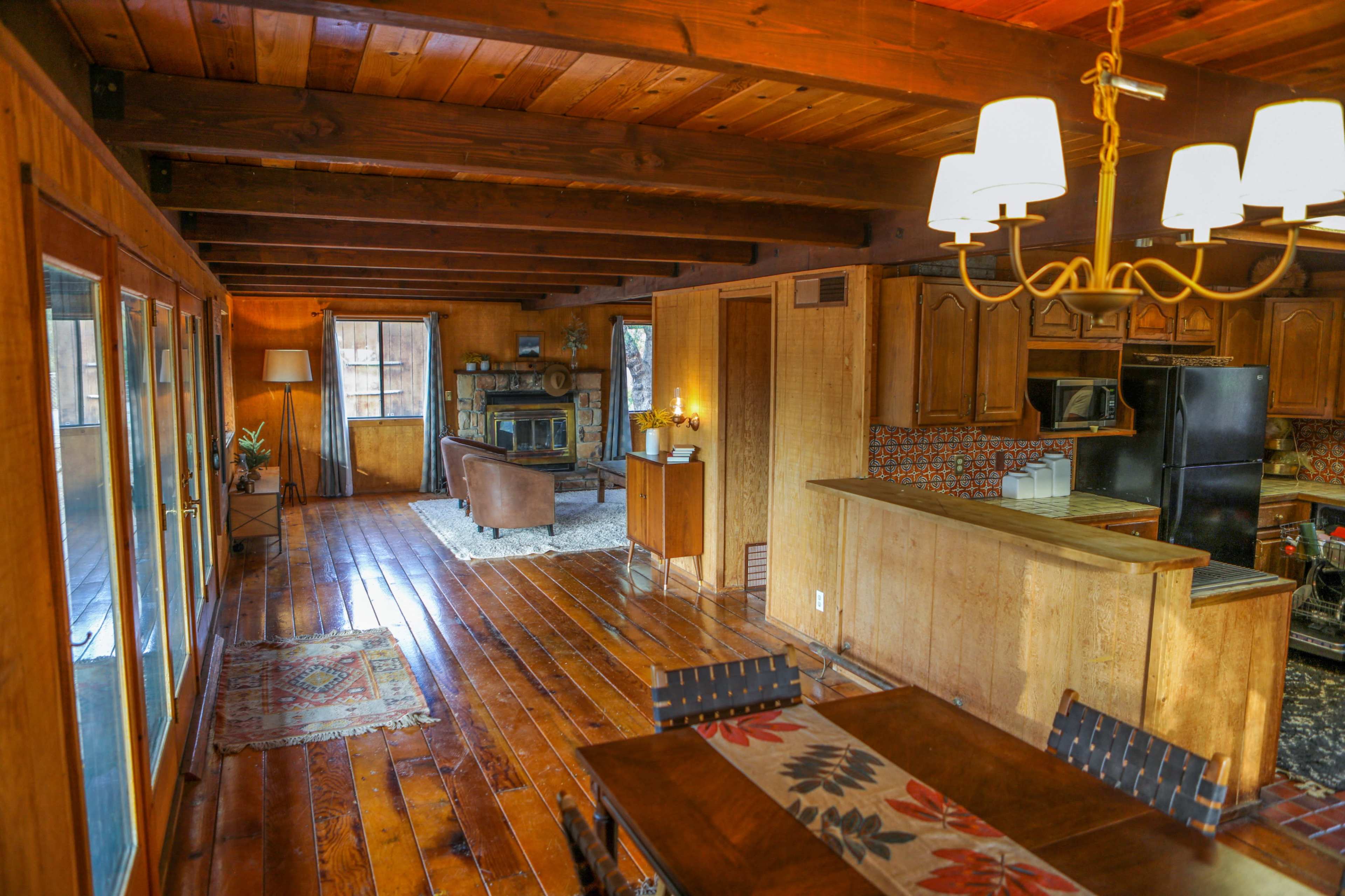 A wooden interior of a cozy living space featuring a kitchen area, dining table, and a seating area with a couch and a fireplace.