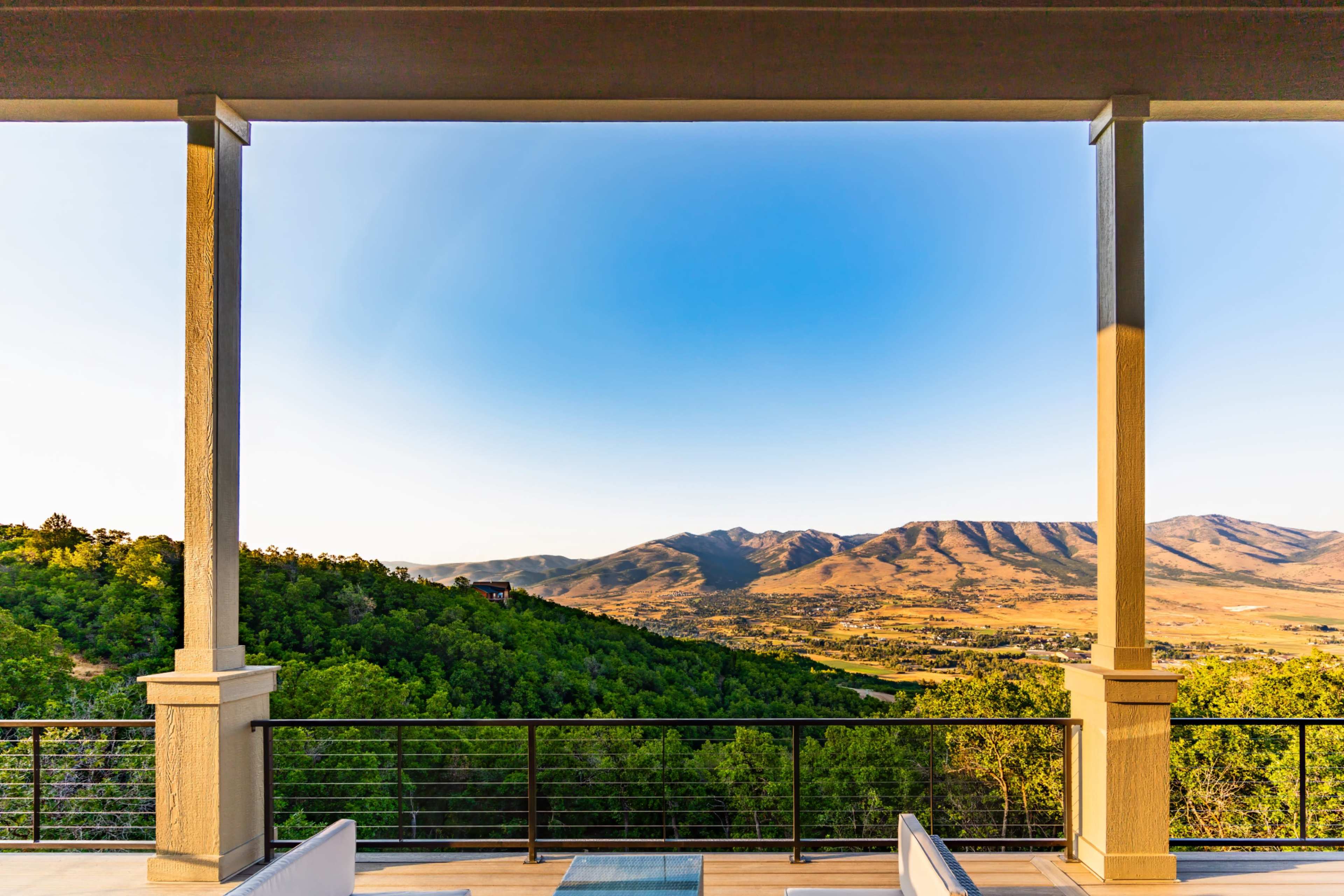 The image shows a spacious balcony with a railing, overlooking a green valley and mountain range under a clear blue sky.