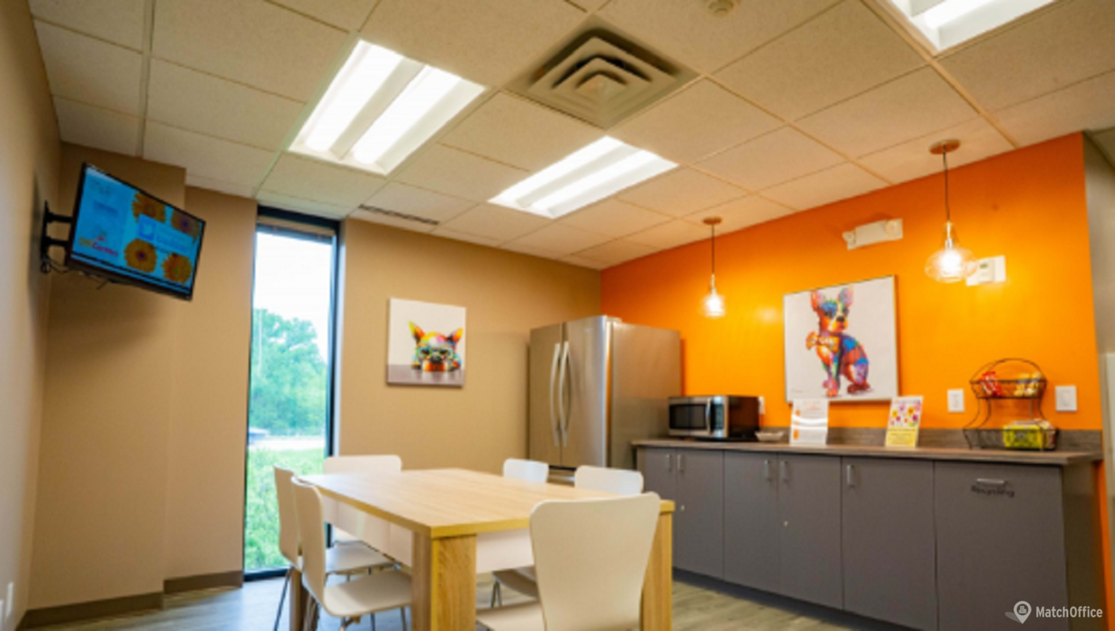 The image shows a modern kitchenette with a large table, a refrigerator, a microwave, and colorful artwork on the walls.