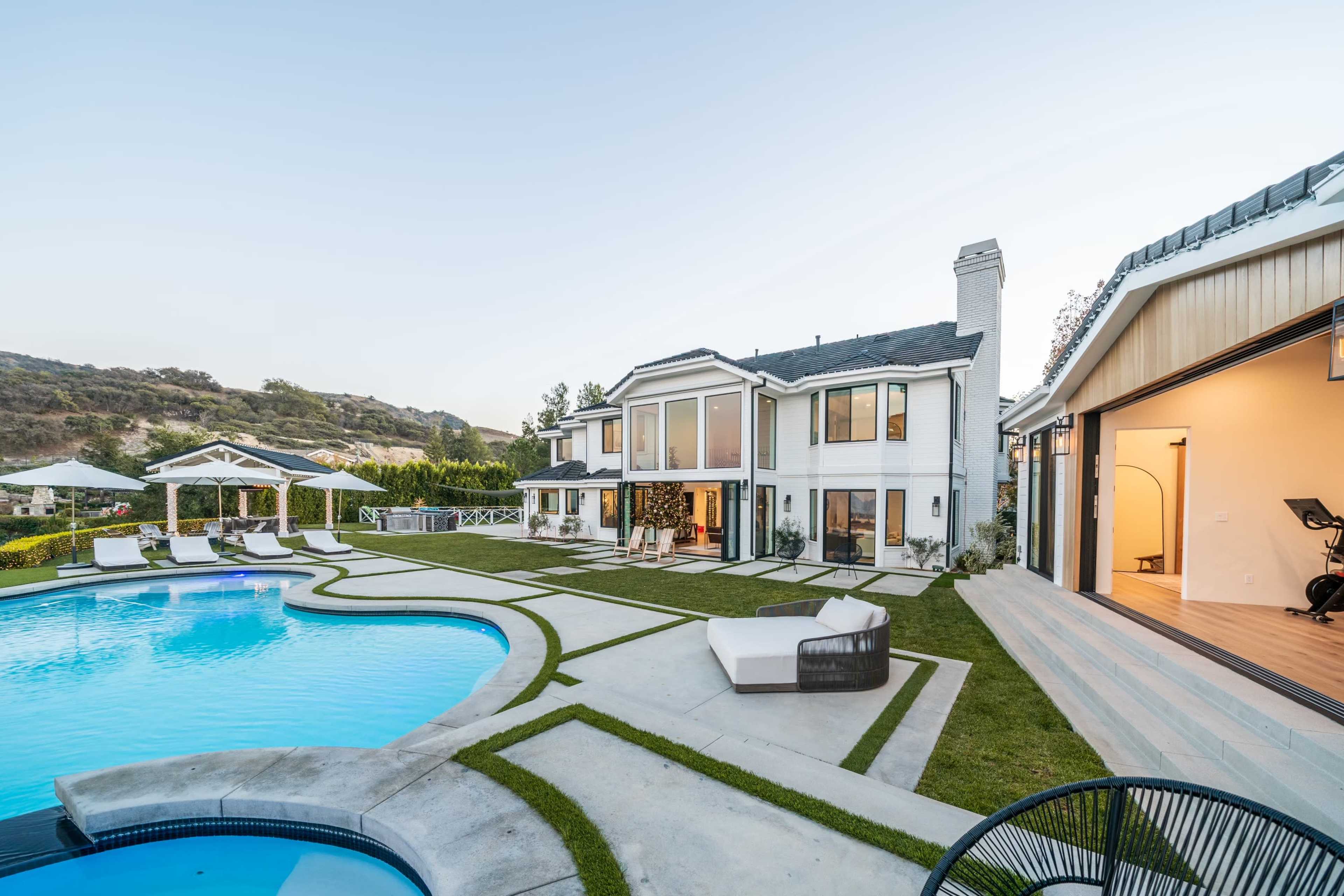A modern two-story house with large windows, a swimming pool, and a landscaped yard featuring lounge chairs and fitness equipment in a garage.