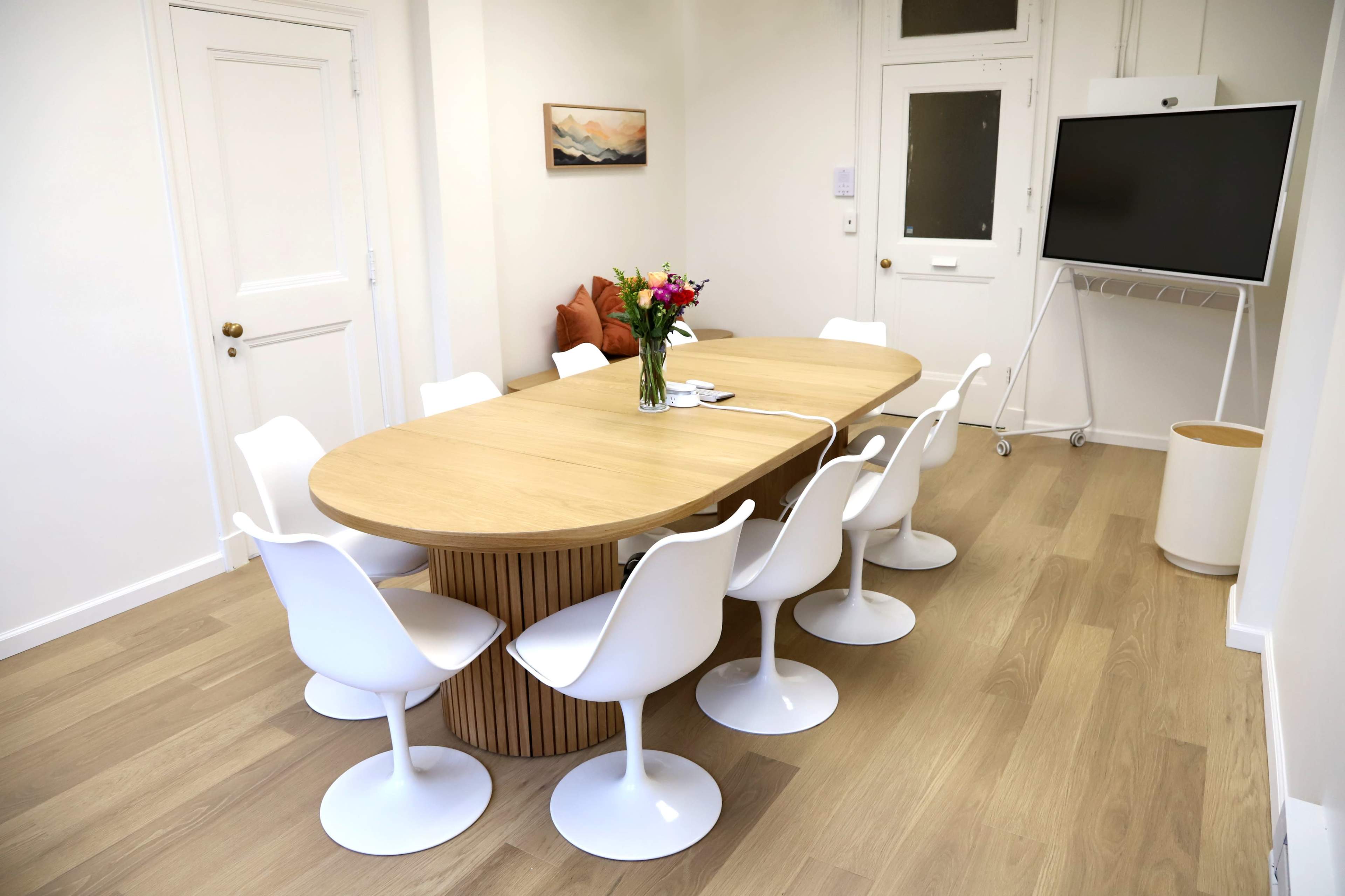 A modern meeting room features a large, oval wooden table surrounded by white, curved chairs, with a vase of flowers at the center.