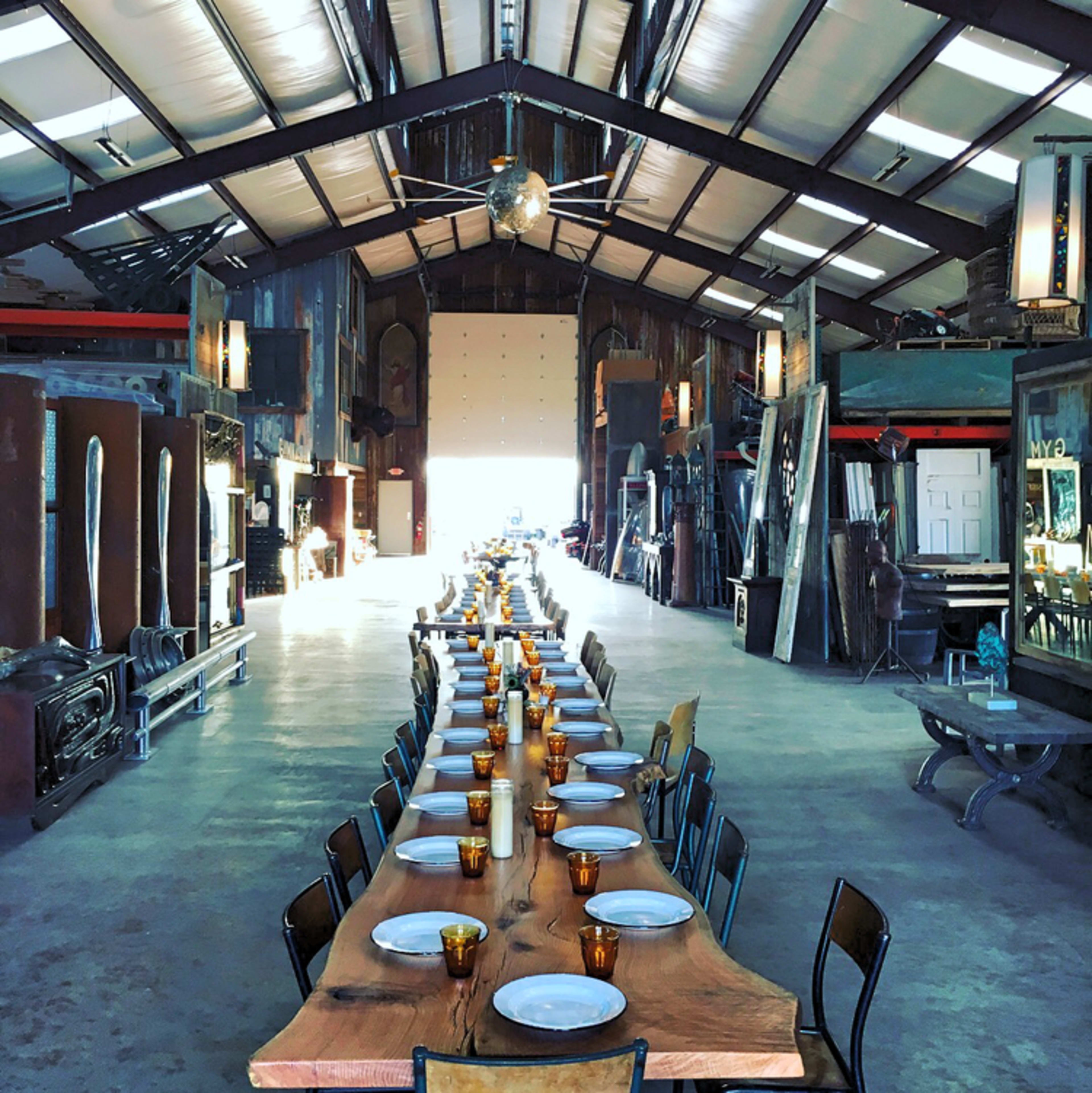 A long, rustic wooden table is set with plates and amber glasses inside a spacious, industrial-style venue with high ceilings and large doors at the far end.