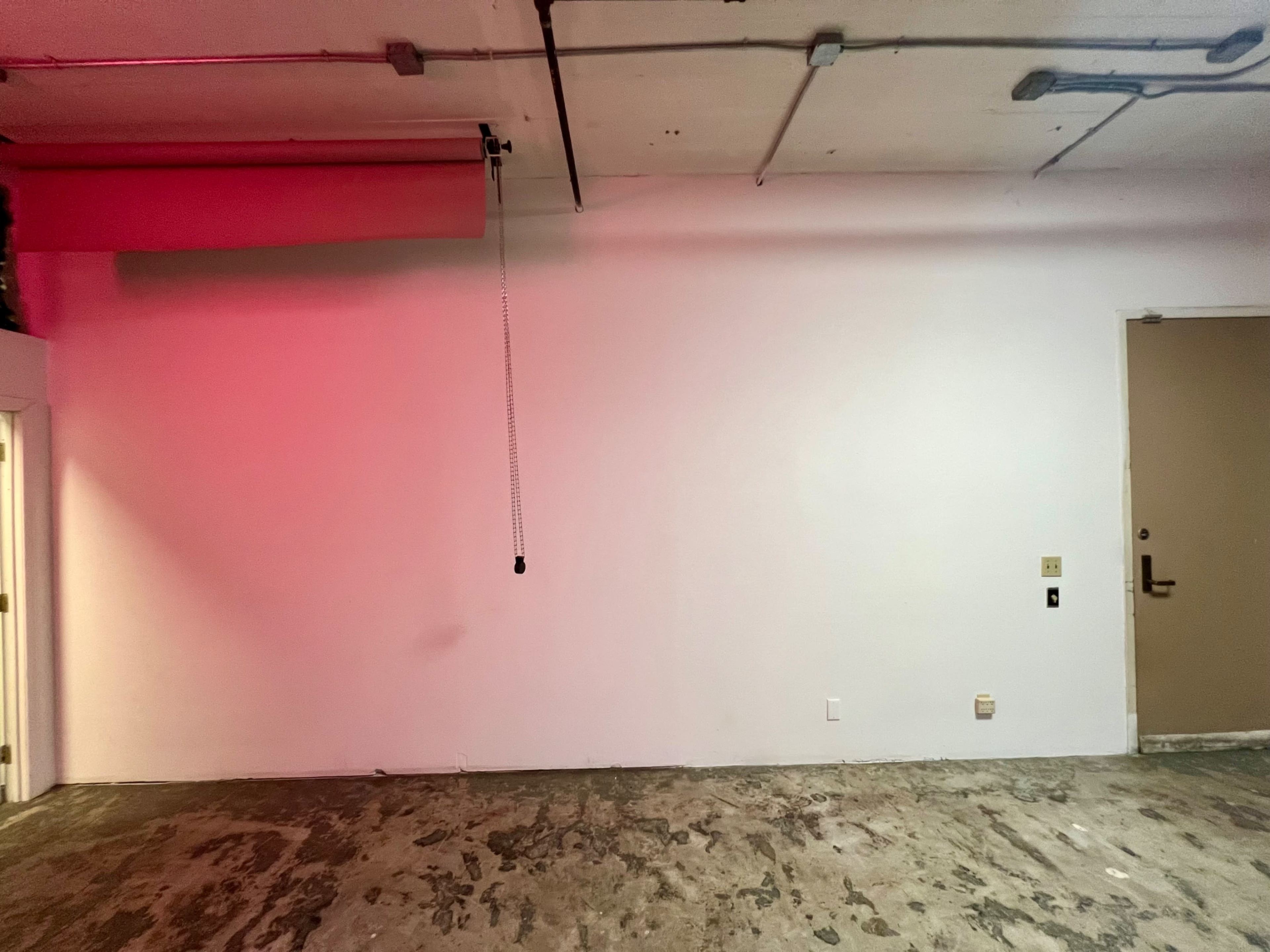 The image shows a vacant room with a concrete floor and a blank white wall, featuring a red light on the left side and a closed door on the right.