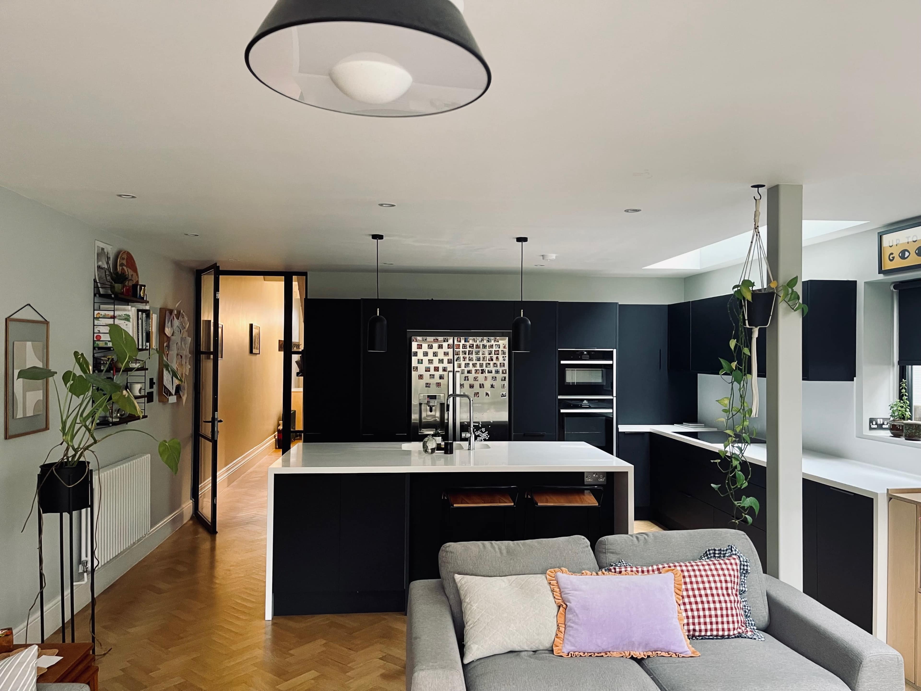 The image shows a modern kitchen with dark cabinetry, a central island, and a spacious open layout connected to a living area with a gray sofa.