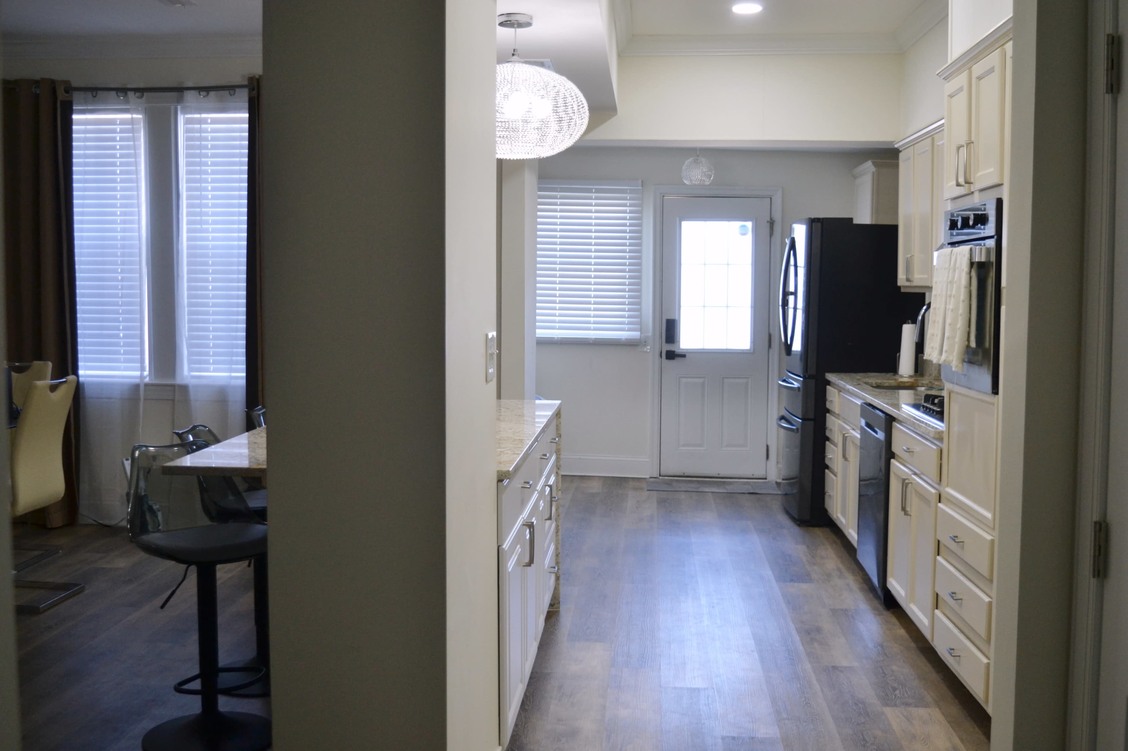 The image shows a modern kitchen with light-colored cabinets, a door leading outside, and a view of a dining area with a glass-top table in the background.