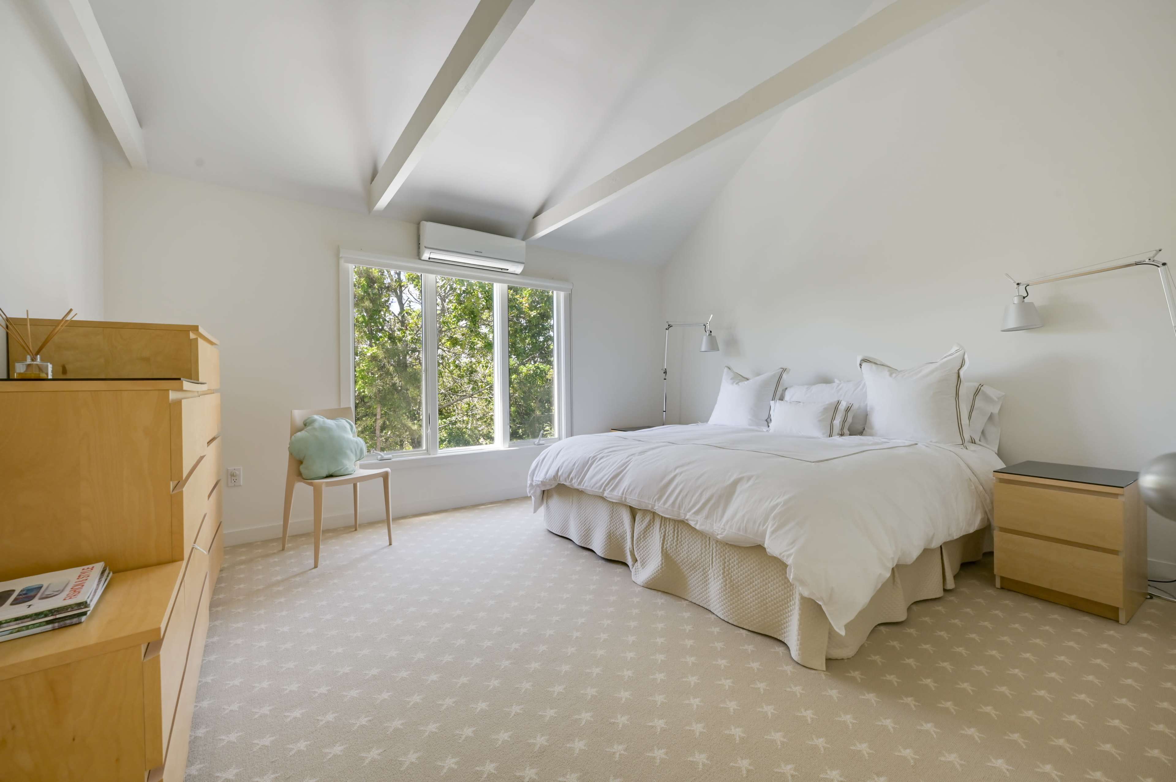 A minimalist bedroom with a large bed, a pair of bedside tables, a wooden dresser, and a chair, all set against white walls and a carpeted floor.