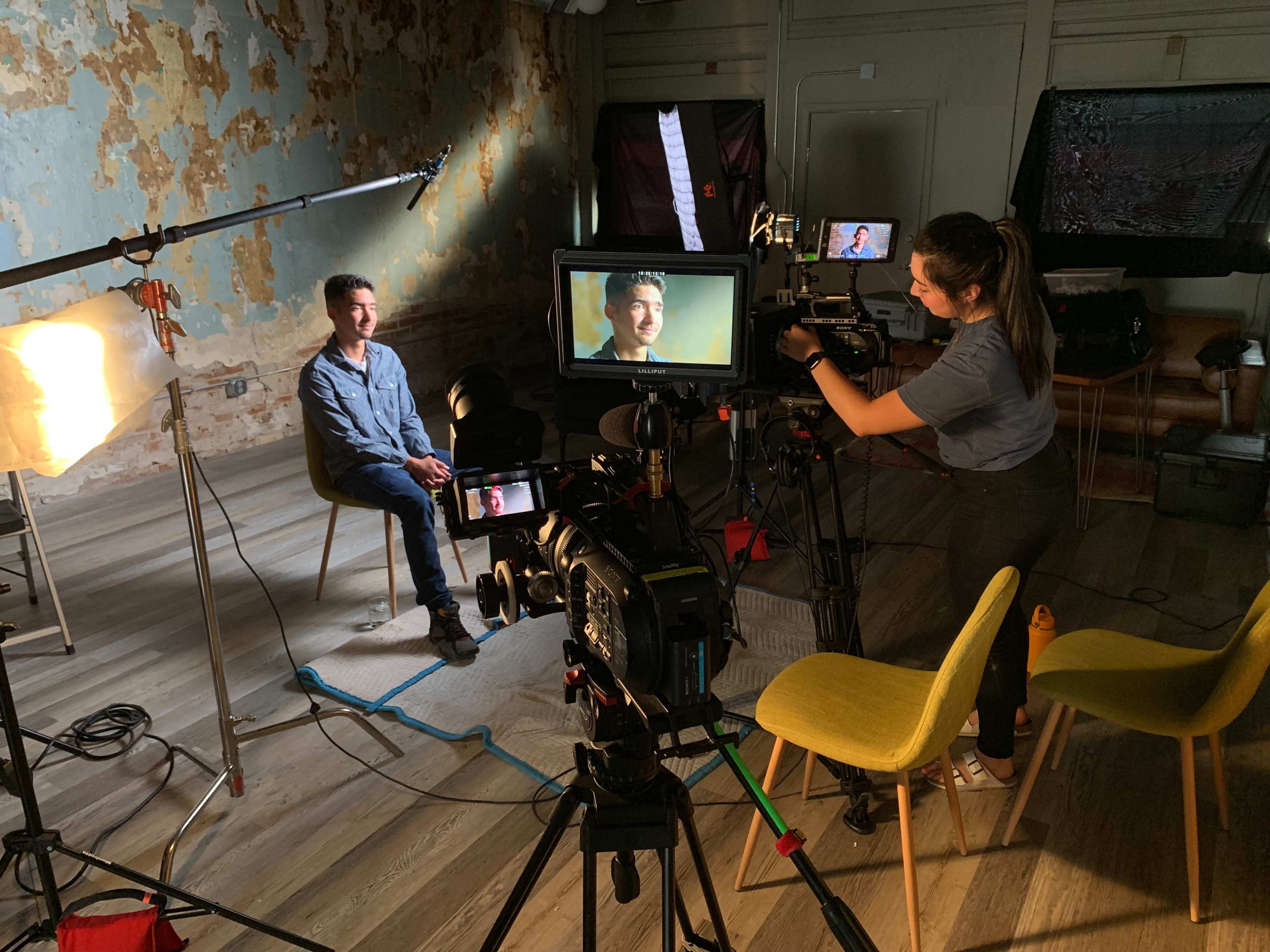 A person is being filmed in a seated interview setup, with cameras, lights, and a crew member adjusting equipment in a room with a distressed wall.