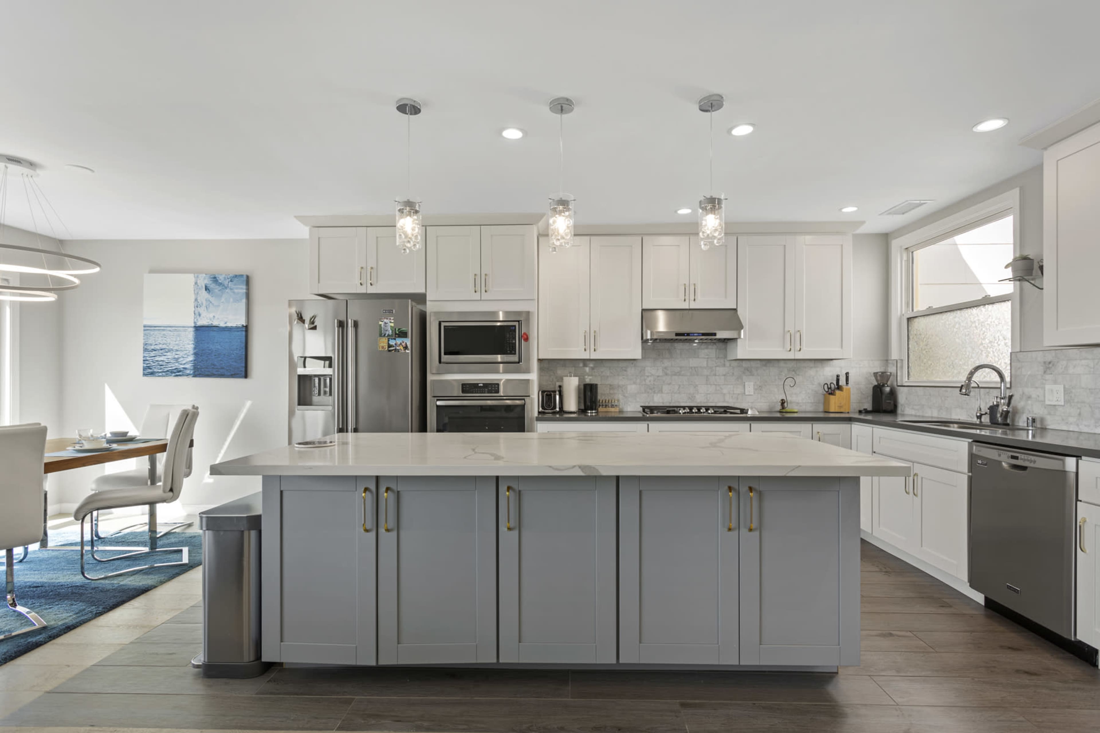 A modern kitchen with white cabinets, a large gray island, stainless steel appliances, and a stylish dining area.
