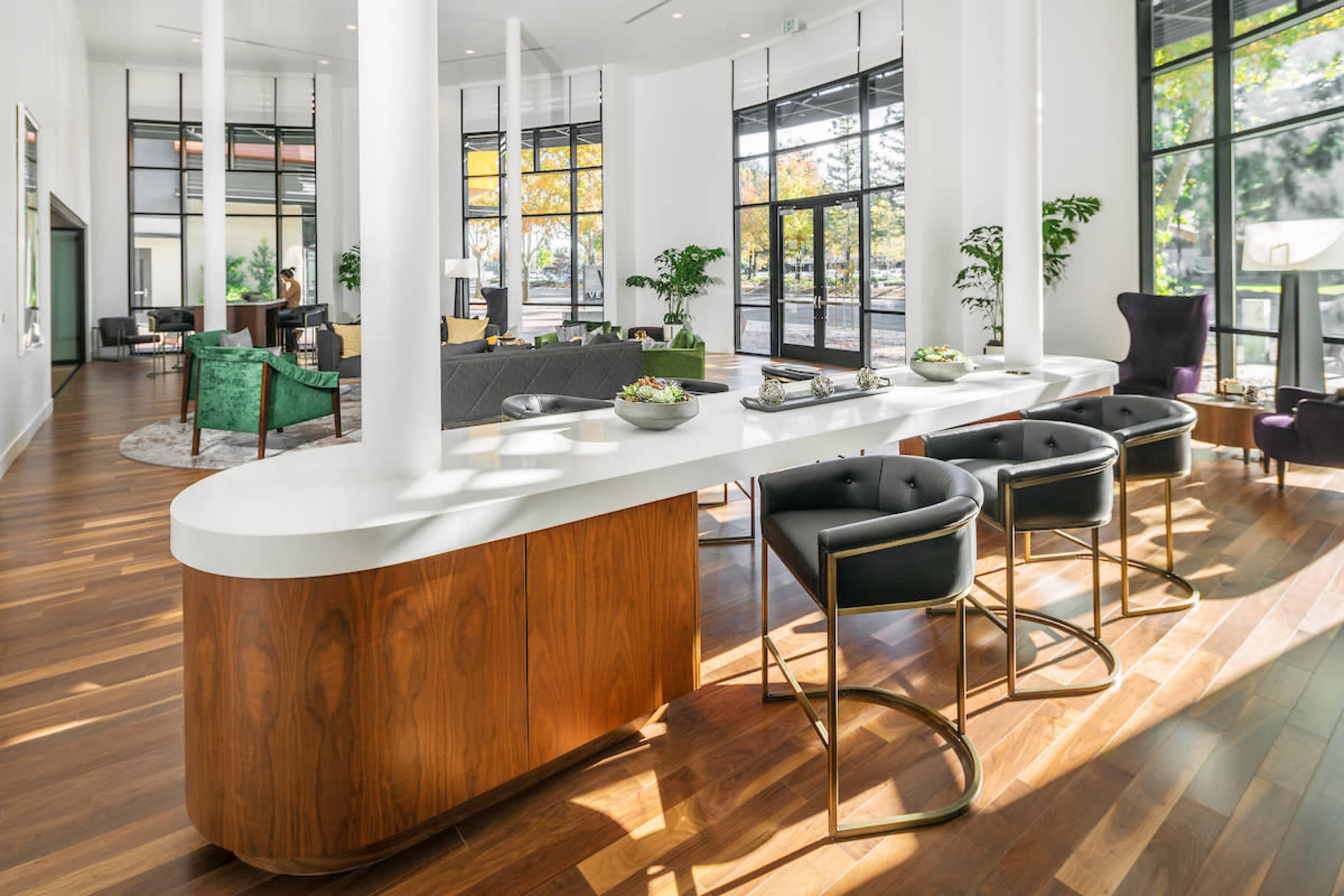 A spacious modern interior features a white countertop bar with black stools, surrounded by large windows and contemporary furniture.