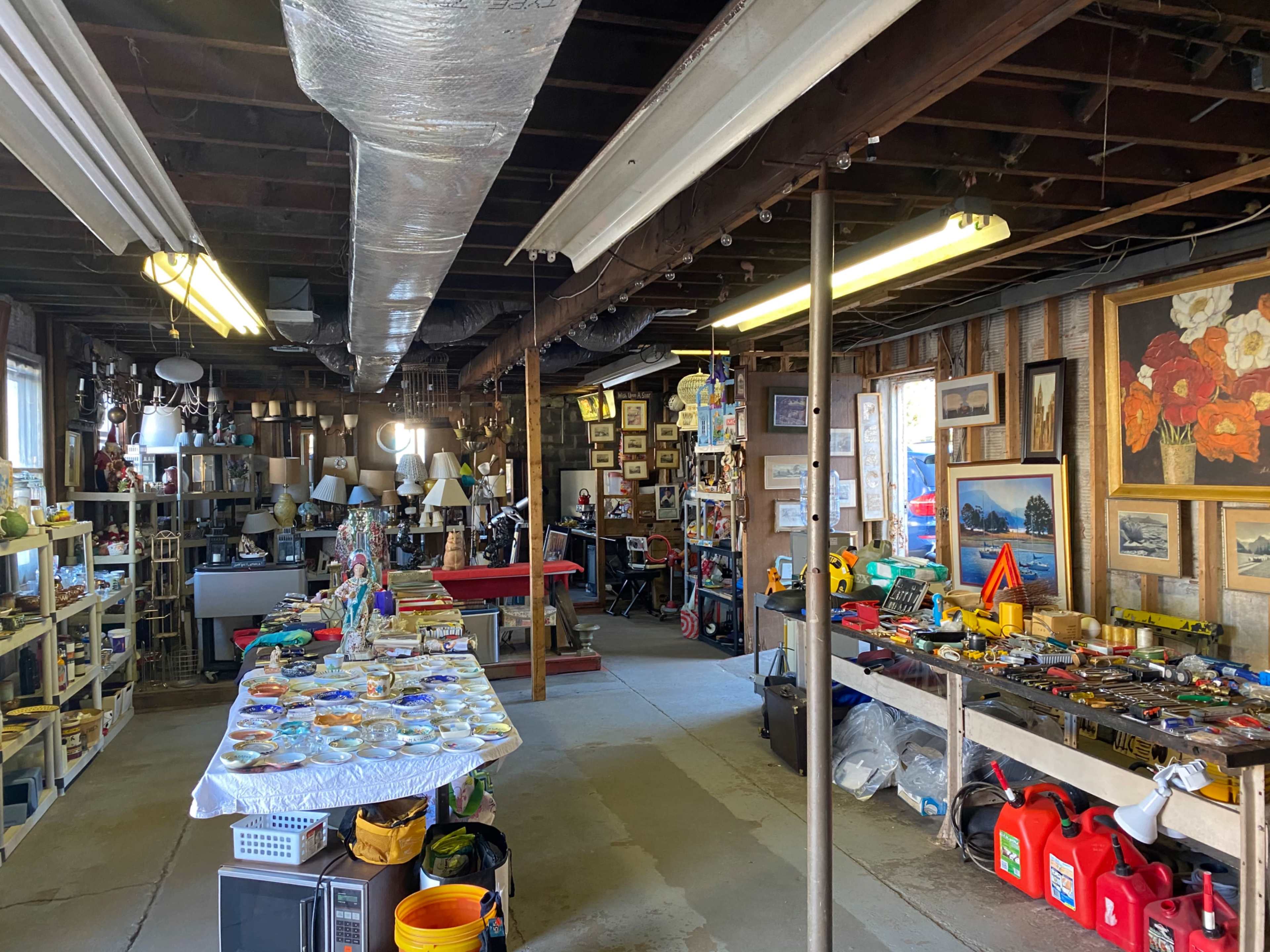 A spacious garage filled with shelves of various items, including tools, lamps, paintings, and a table displaying decorative objects.
