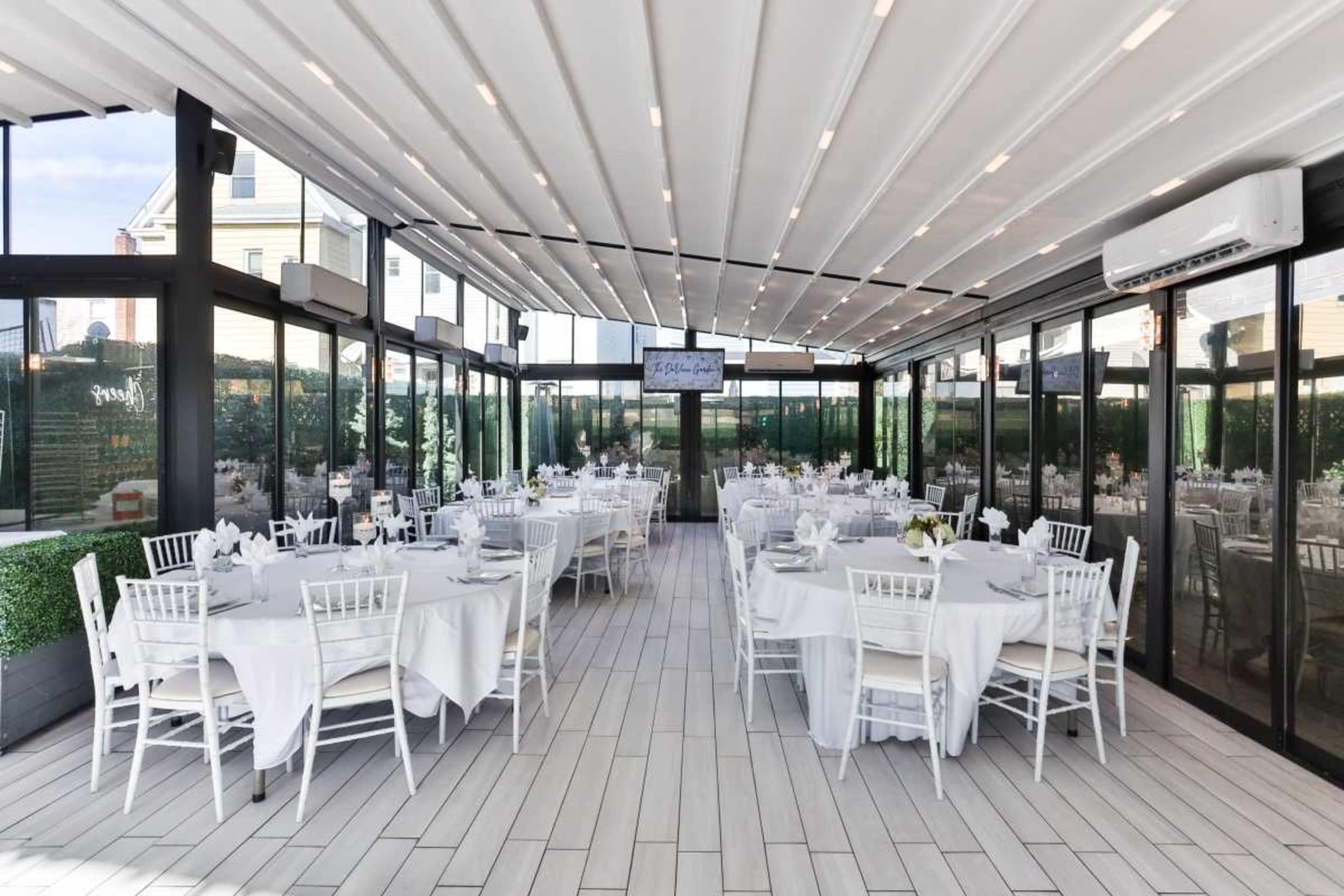 The image shows a bright, modern dining space with round tables set for a meal, surrounded by large glass walls and a white ceiling.