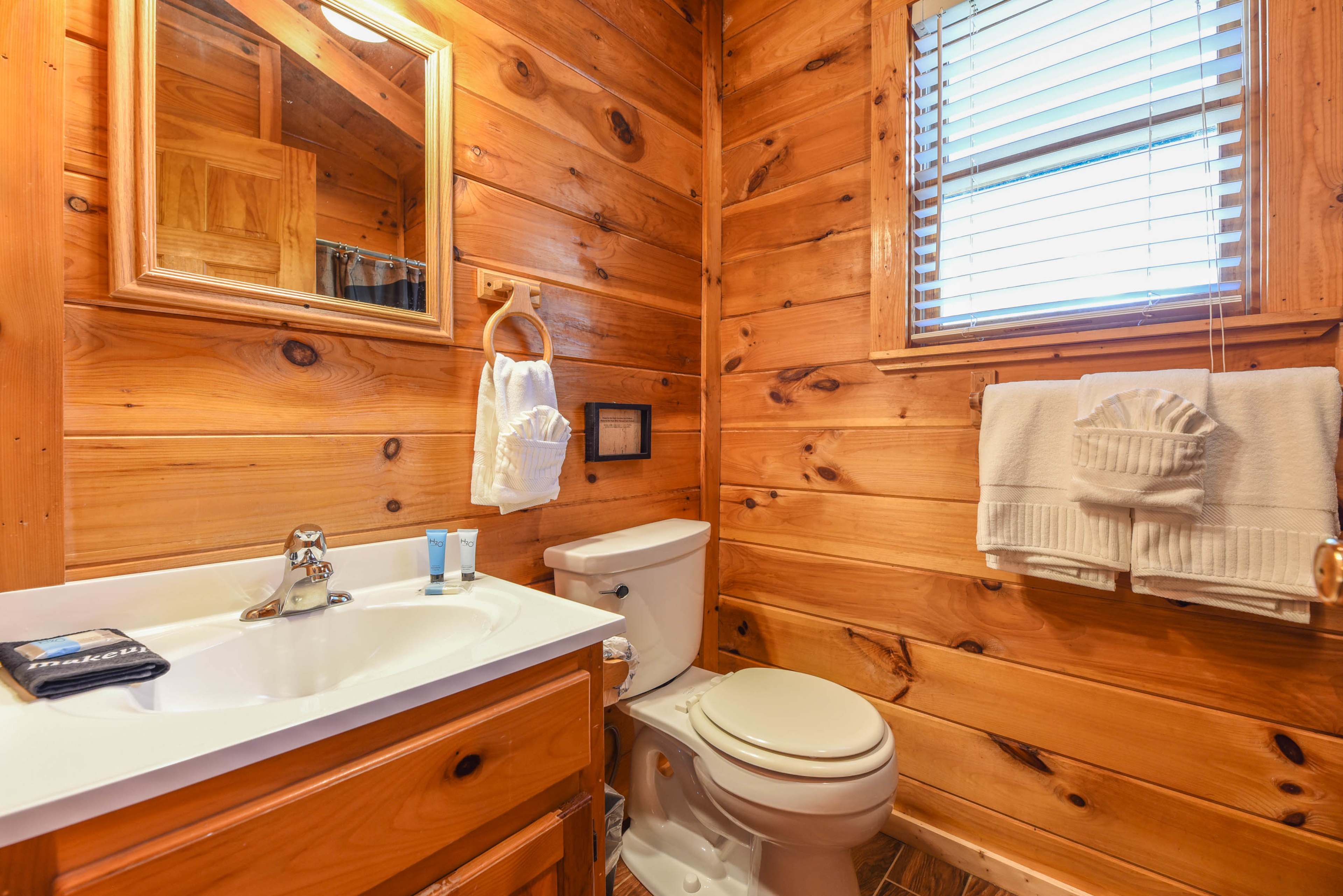 The image shows a bathroom with wooden walls, a sink with a mirror above, a toilet, and neatly arranged towels.