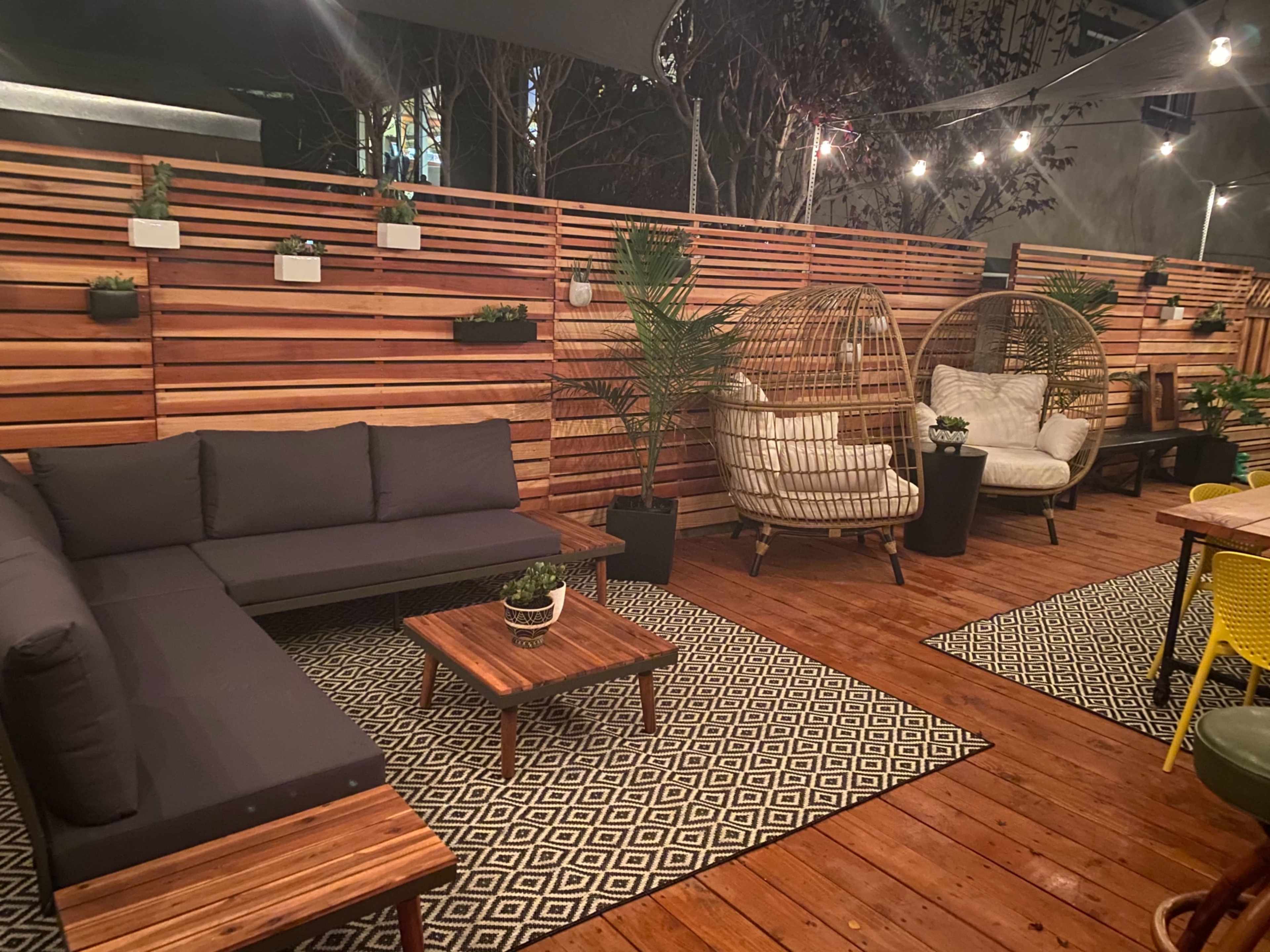 A cozy outdoor seating area with wood paneling, plush seating, and decorative plants, illuminated by string lights.