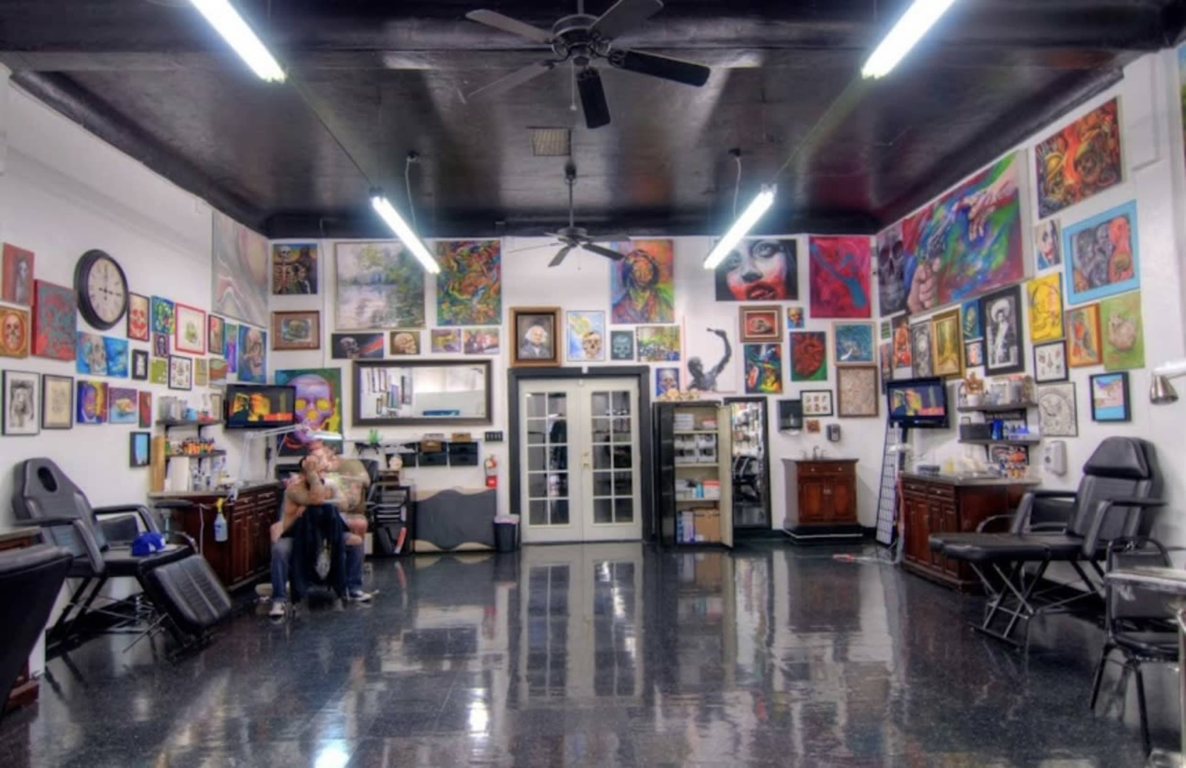 The image shows an art-filled tattoo parlor with various artworks displayed on the walls and tattoo stations arranged around the room.