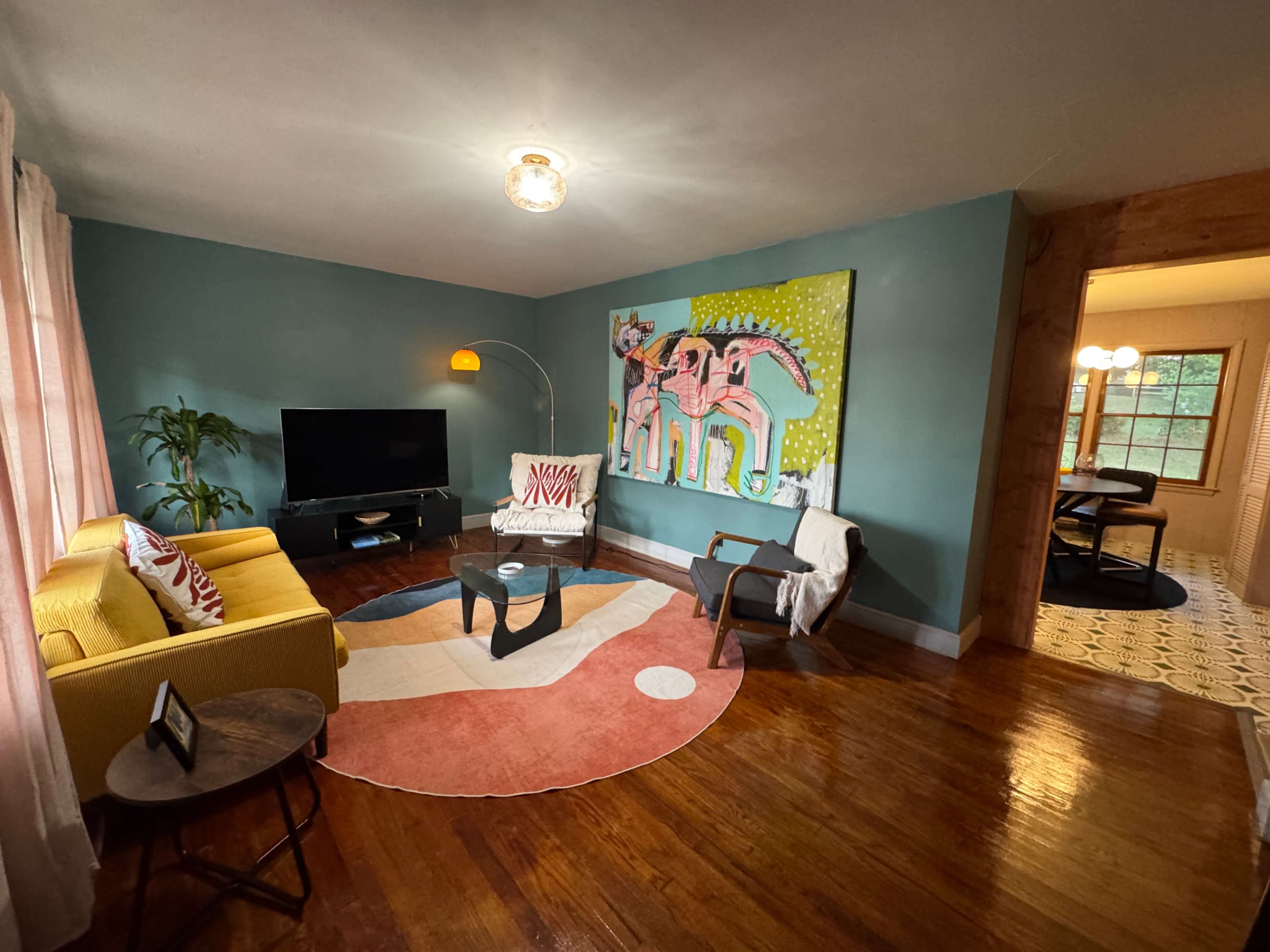 The image shows a colorful living room with a yellow sofa, a large elephant painting on the wall, and a circular area rug.