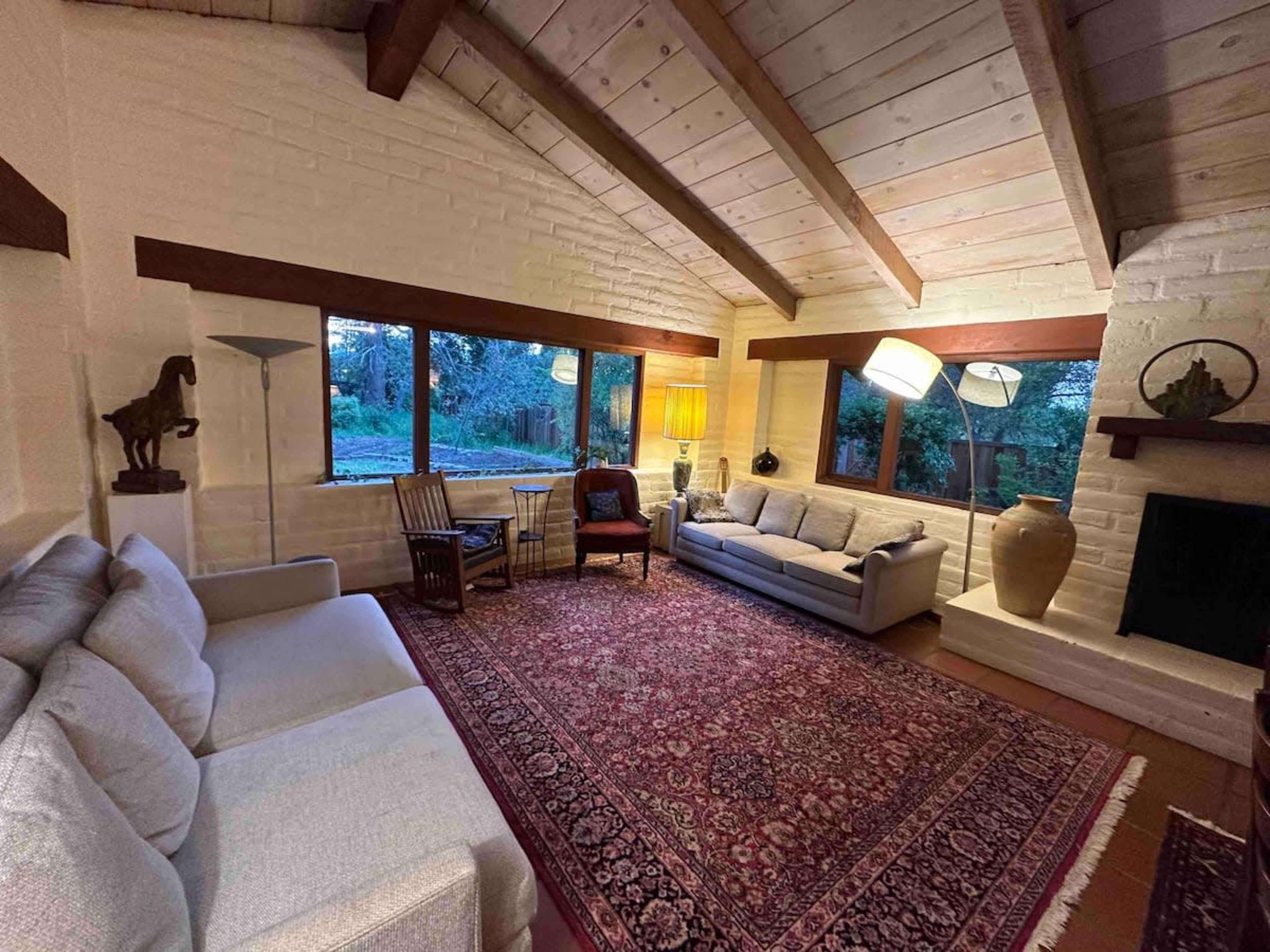The scene displays a cozy living room featuring a gray sofa, wooden chairs, a large area rug, and large windows overlooking a garden.