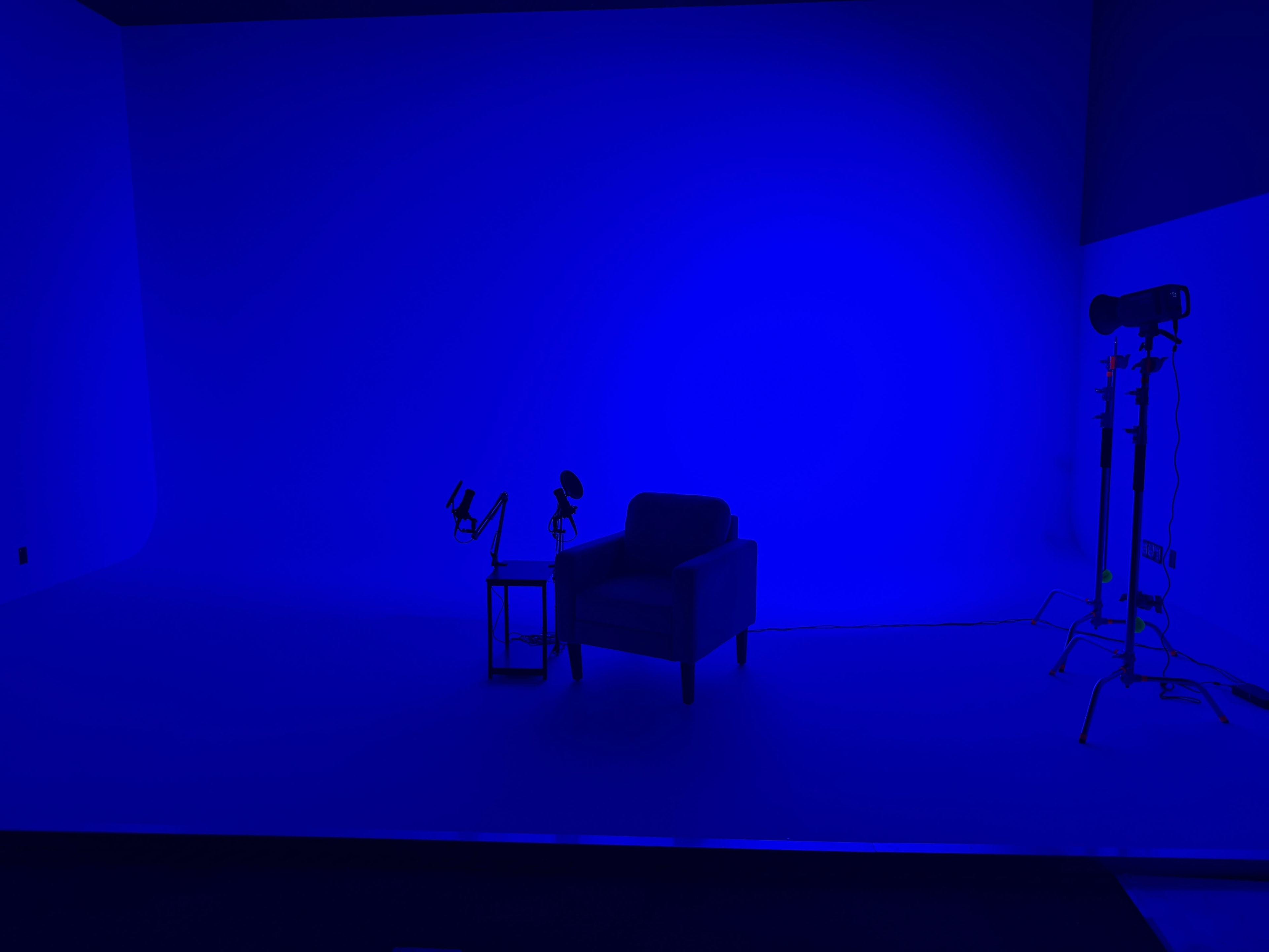 The image shows a blue-lit studio space featuring a chair, a small table, and lighting equipment.
