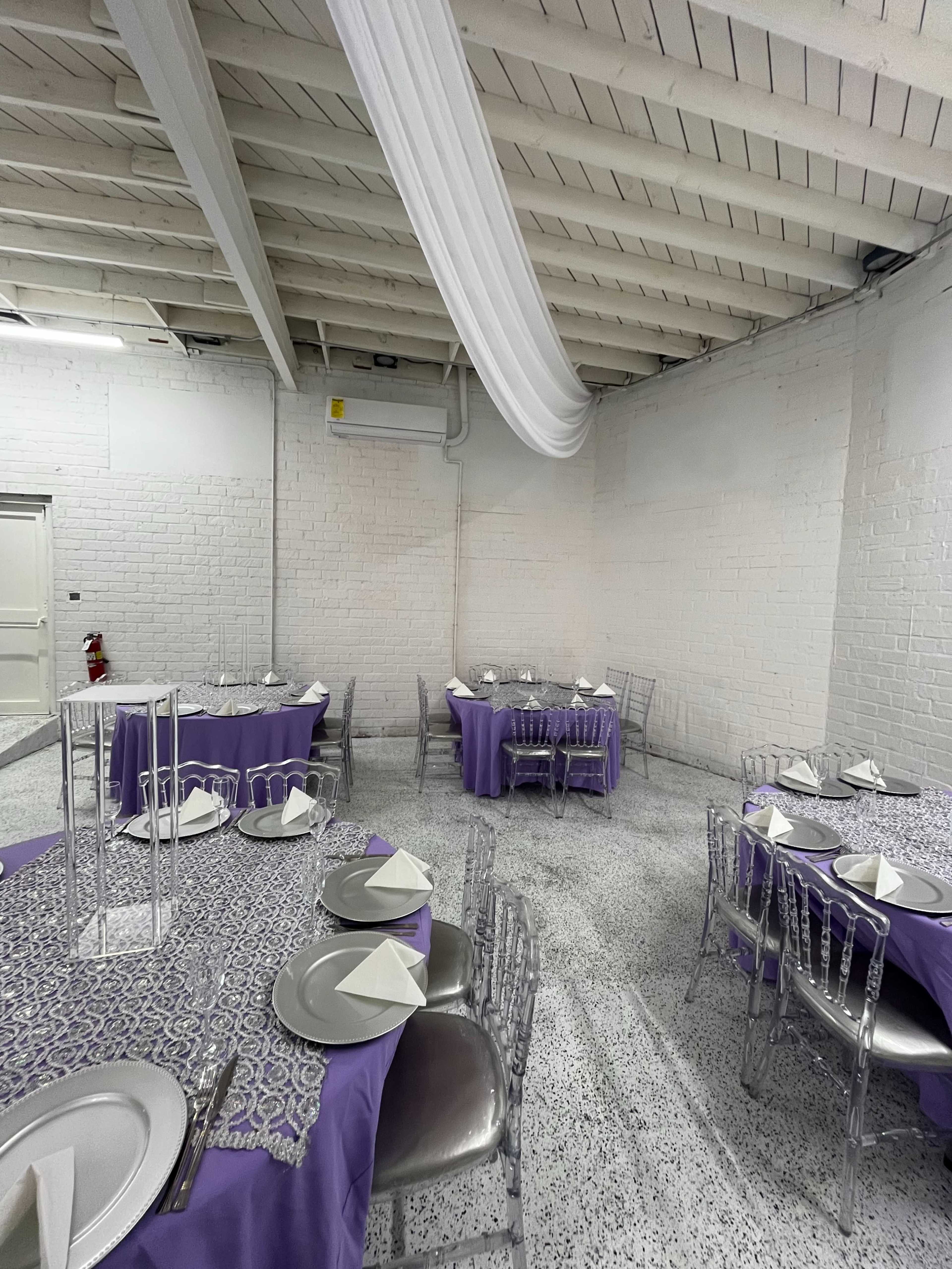 The image shows a banquet hall featuring round tables covered with purple tablecloths and set with silver plates and napkins, surrounded by metal chiavari chairs.