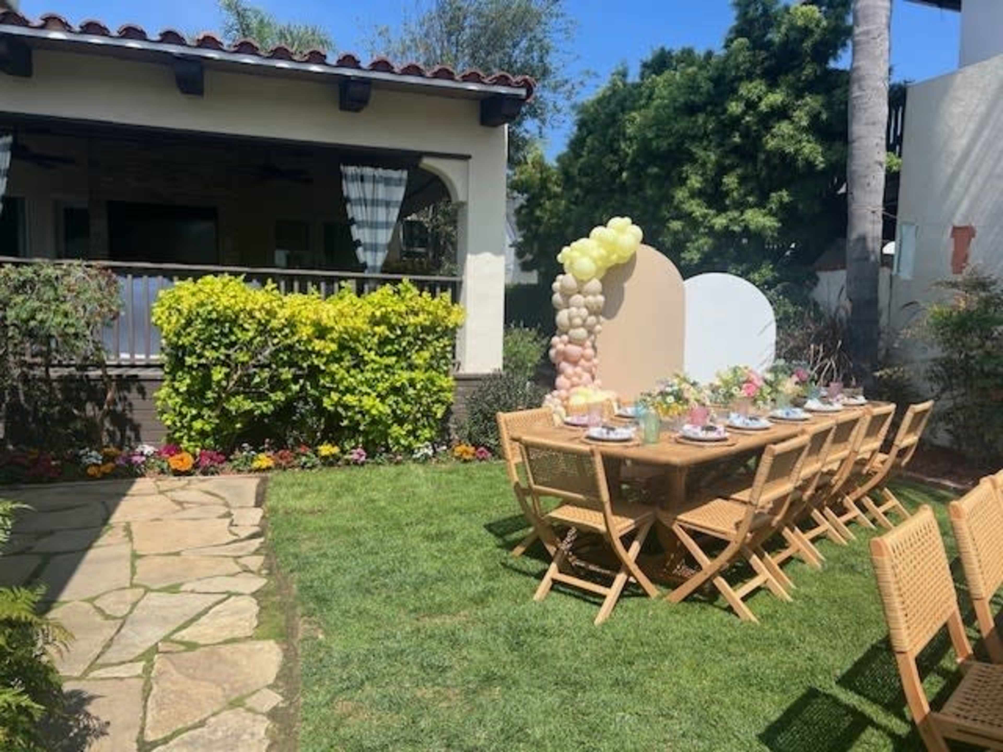 A long, wooden table set for a meal is placed on a grassy area surrounded by greenery and colorful flowers, with a house in the background.