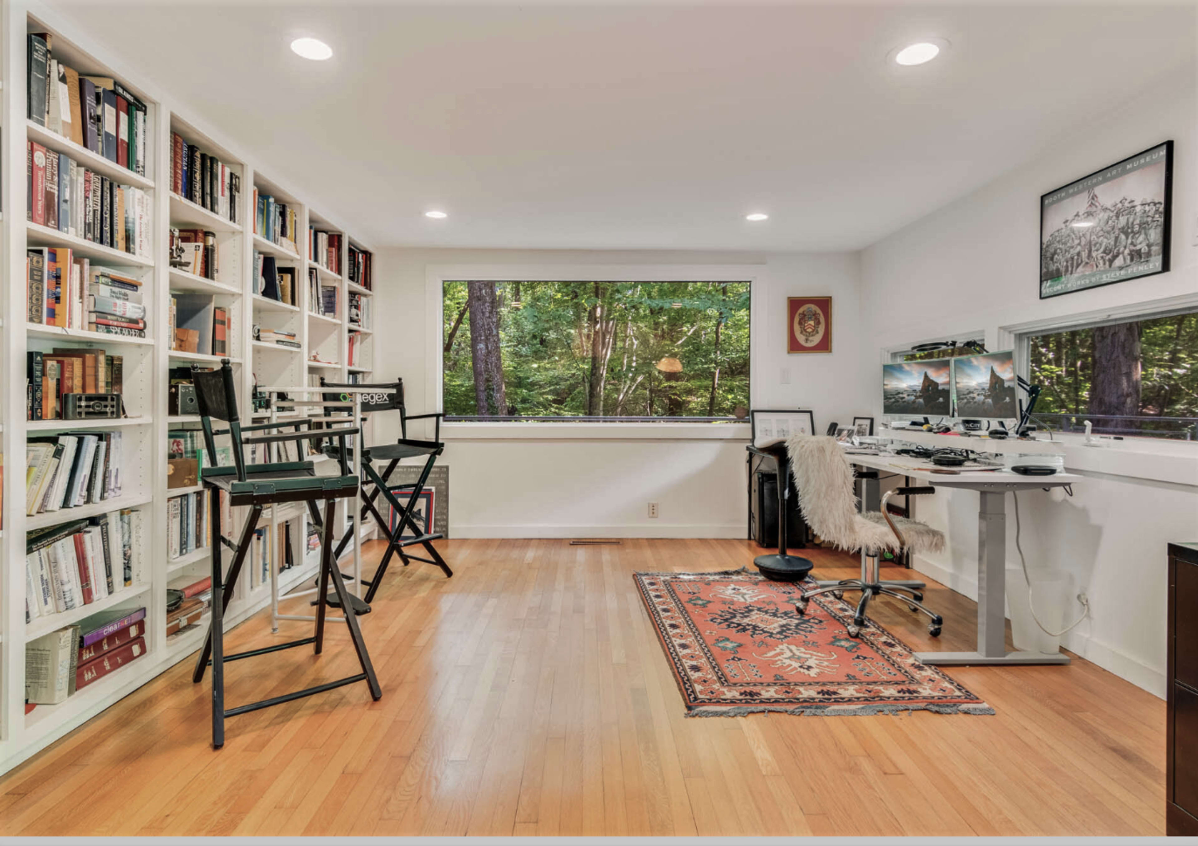 The image shows a bright office space featuring a wall of bookshelves, a desk with a computer, and large windows overlooking a wooded area.