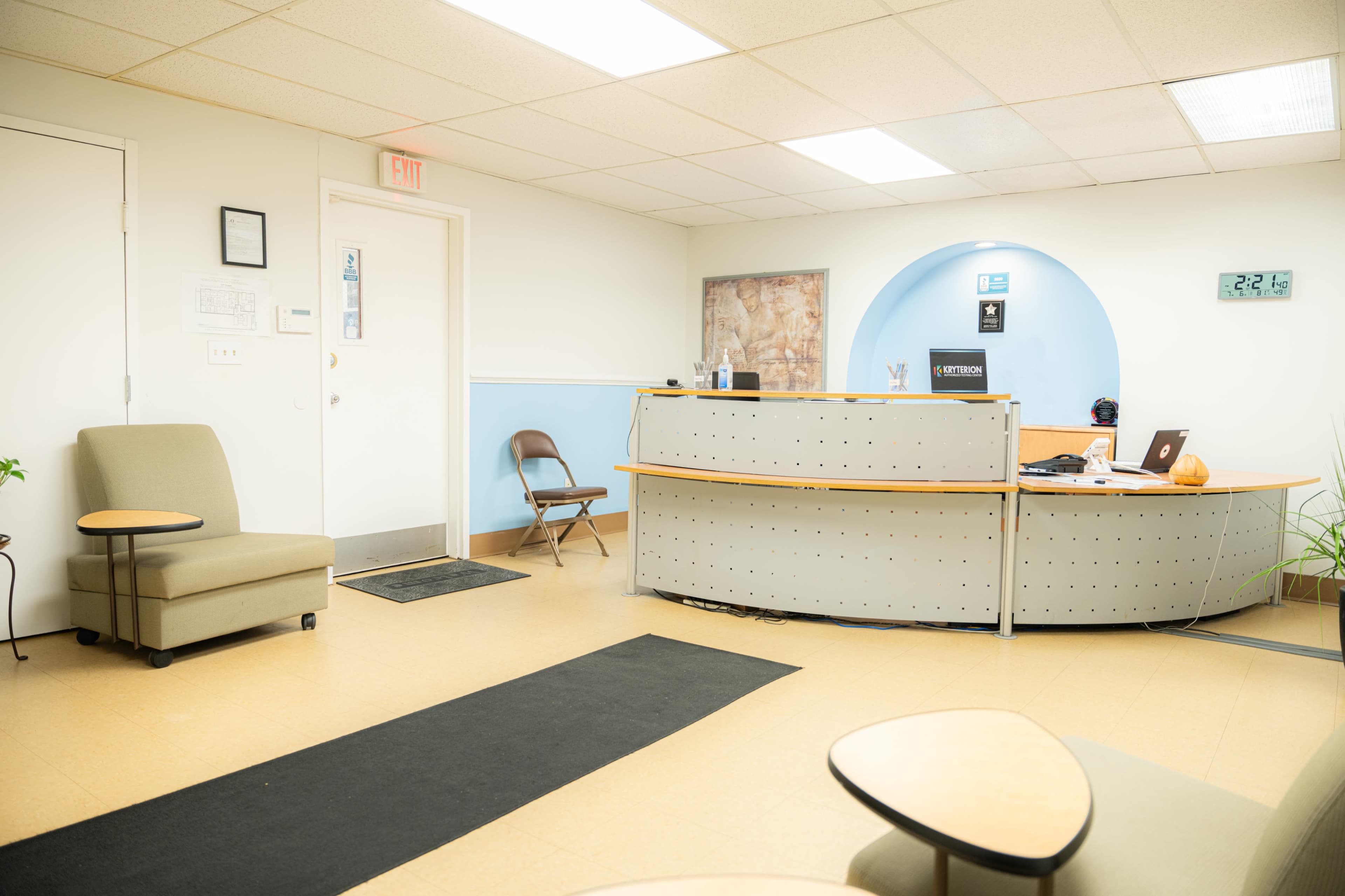 A reception area with a desk, a clock on the wall, and a few chairs arranged around a carpeted floor.