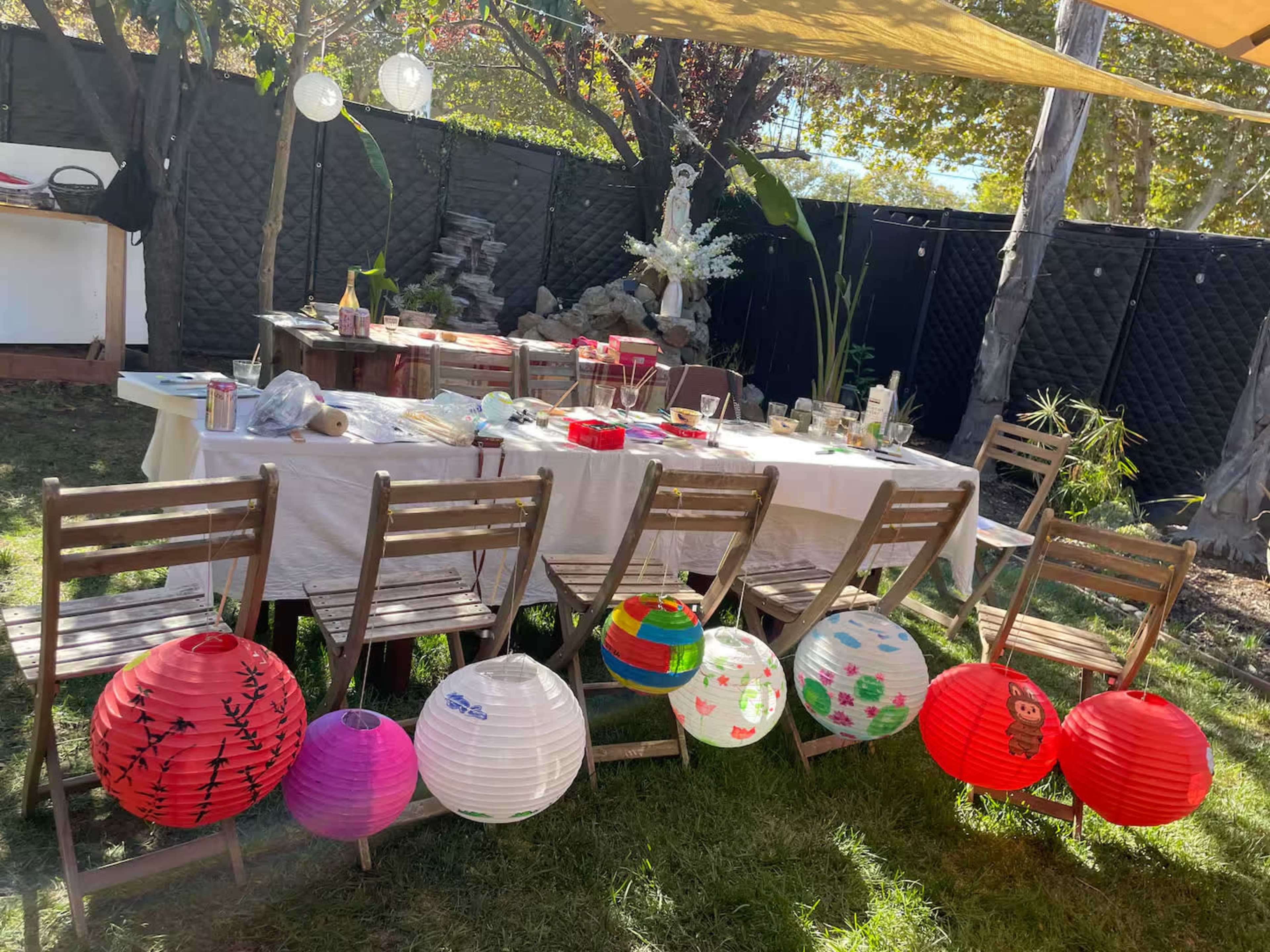 A set of wooden chairs is arranged in a garden, with colorful paper lanterns hanging from them and a table covered with food and decorations nearby.