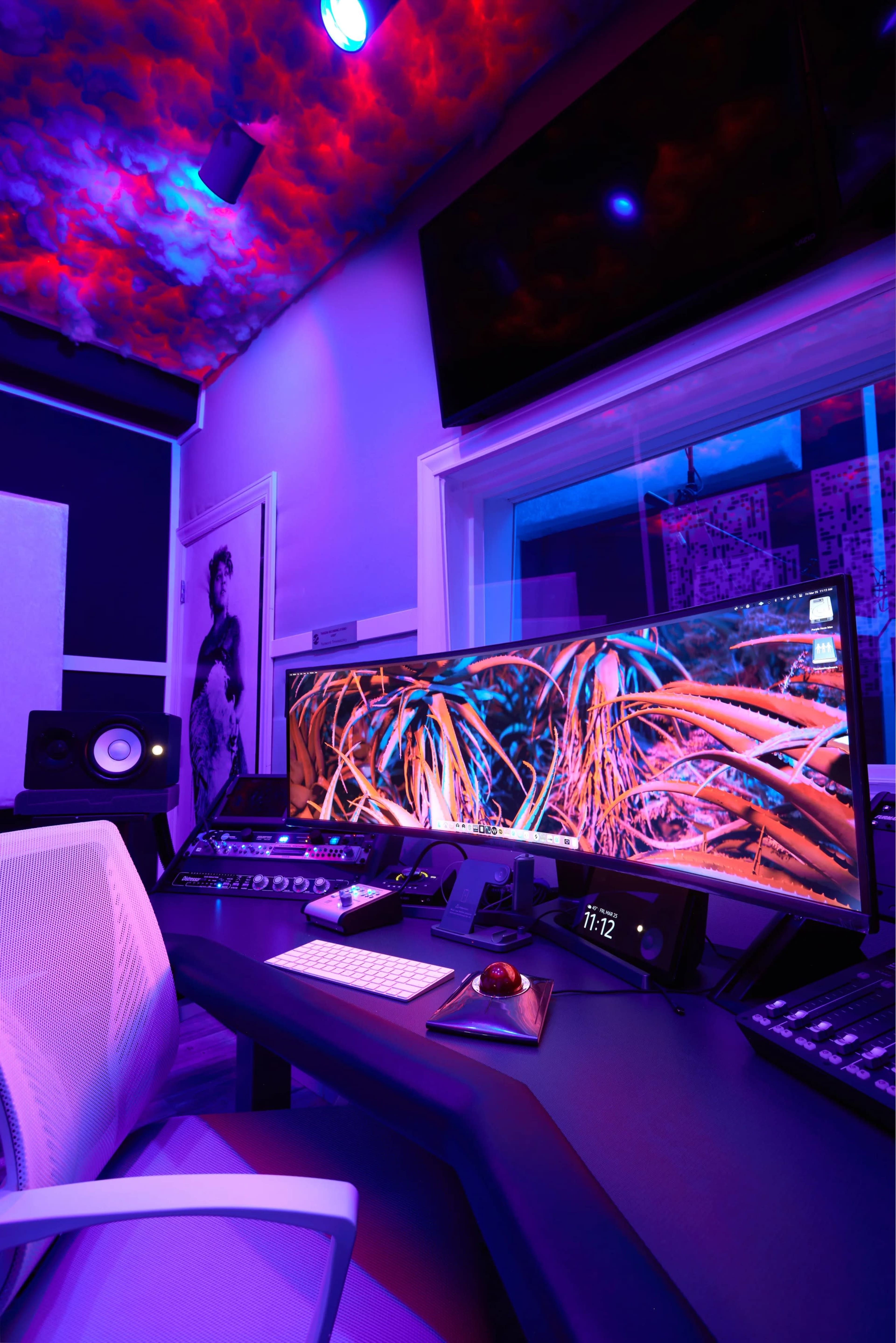 A modern music studio features a large monitor displaying abstract artwork, surrounded by professional audio equipment and colorful lighting.