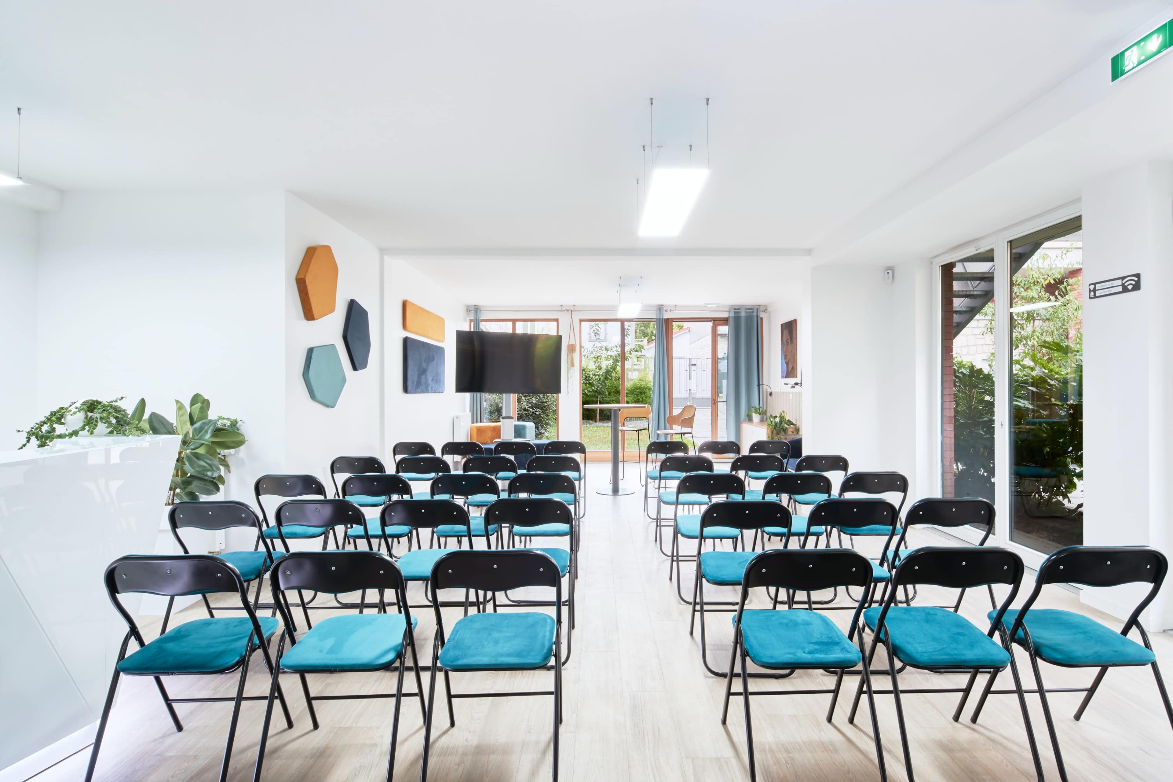 A modern, spacious room arranged with black folding chairs featuring teal cushions, facing a large screen in a setting with large windows and greenery visible outside.
