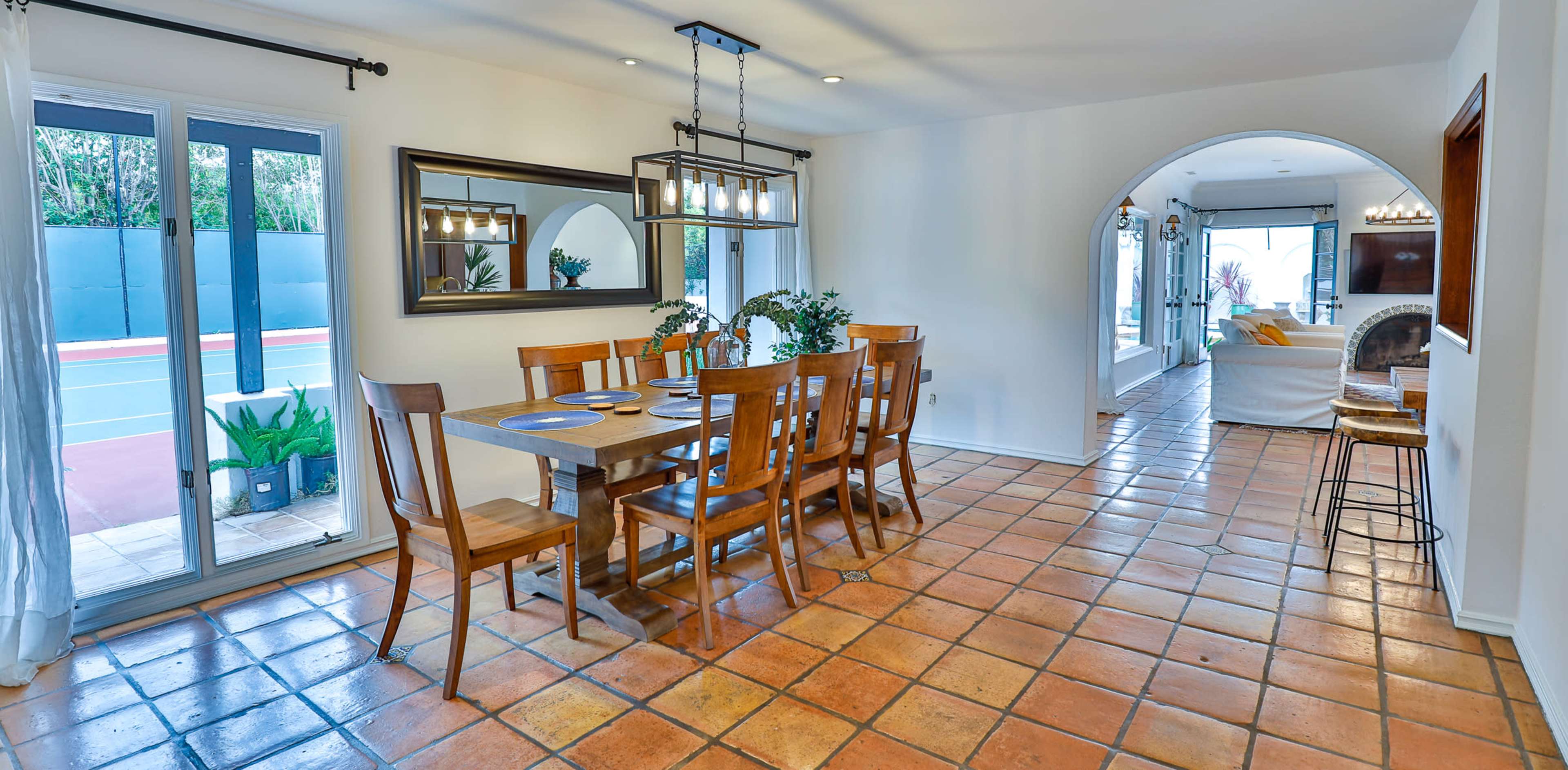 A dining area features a wooden table surrounded by chairs, with a large mirror and pendant lights above, adjacent to a living space with an archway leading outside.