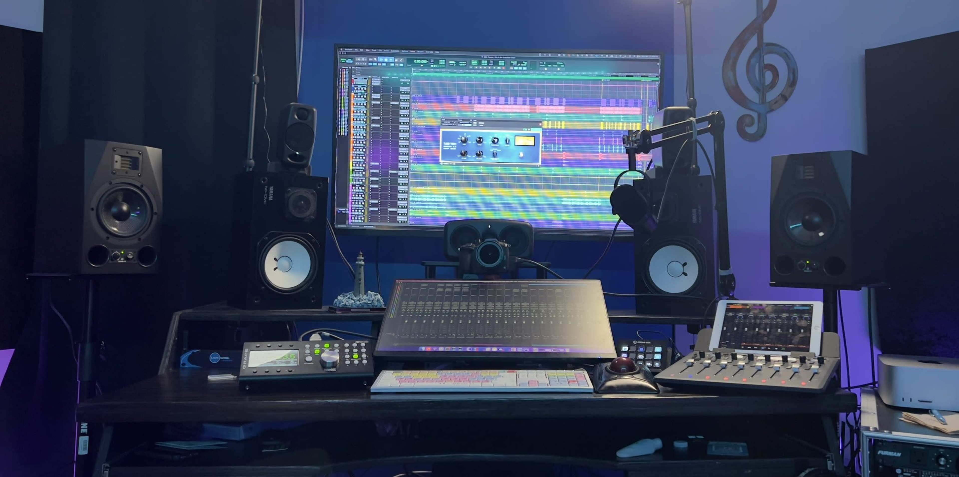 A music production studio setup with a large computer monitor displaying a digital audio workstation, flanked by speakers and various audio equipment on a desk.