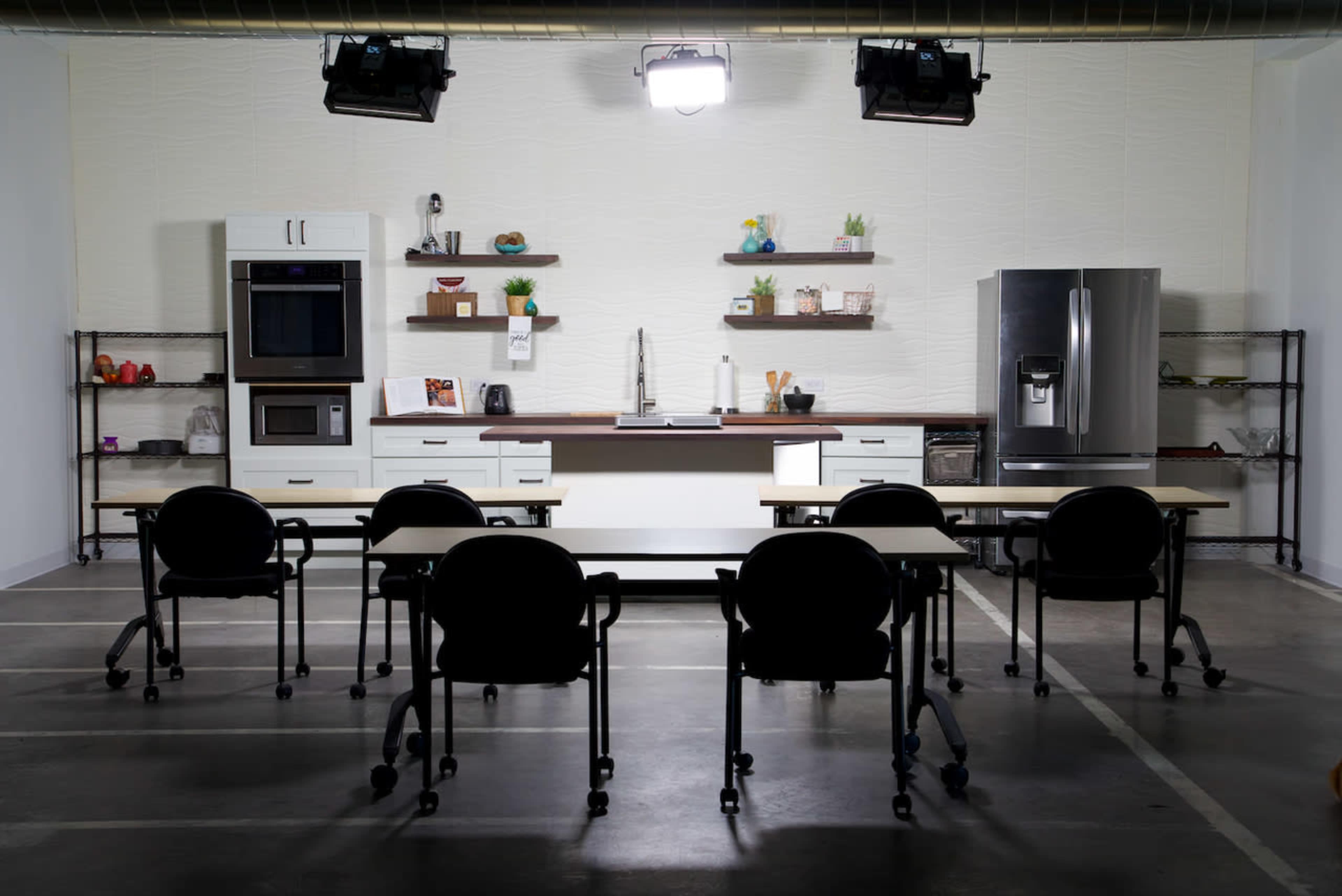 A modern kitchen setup with a stainless steel refrigerator, a wall oven, and a long countertop, accompanied by several tables and chairs arranged in front.