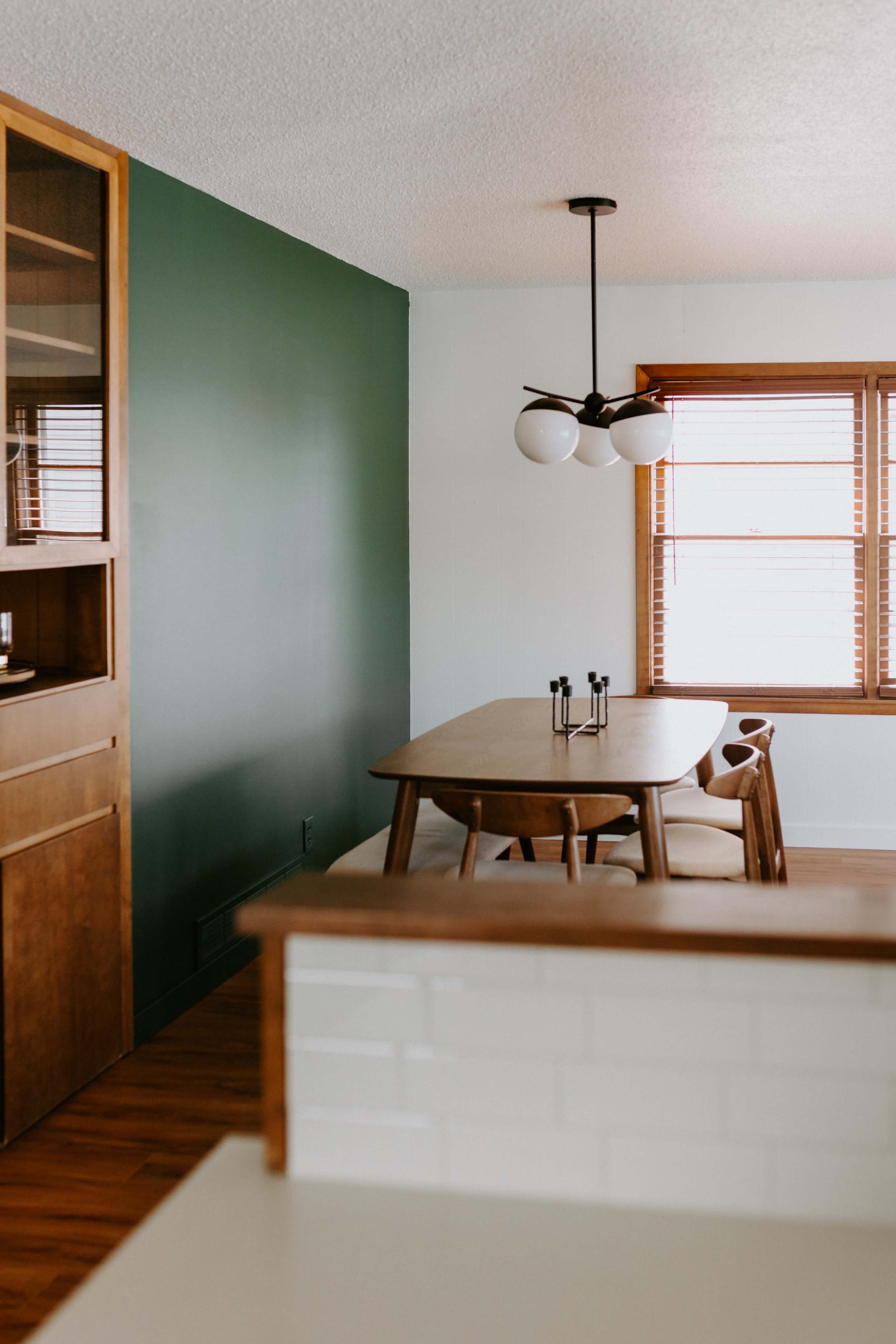 A dining area with a wooden table and chairs under a modern light fixture, situated between a green wall and a window with blinds.