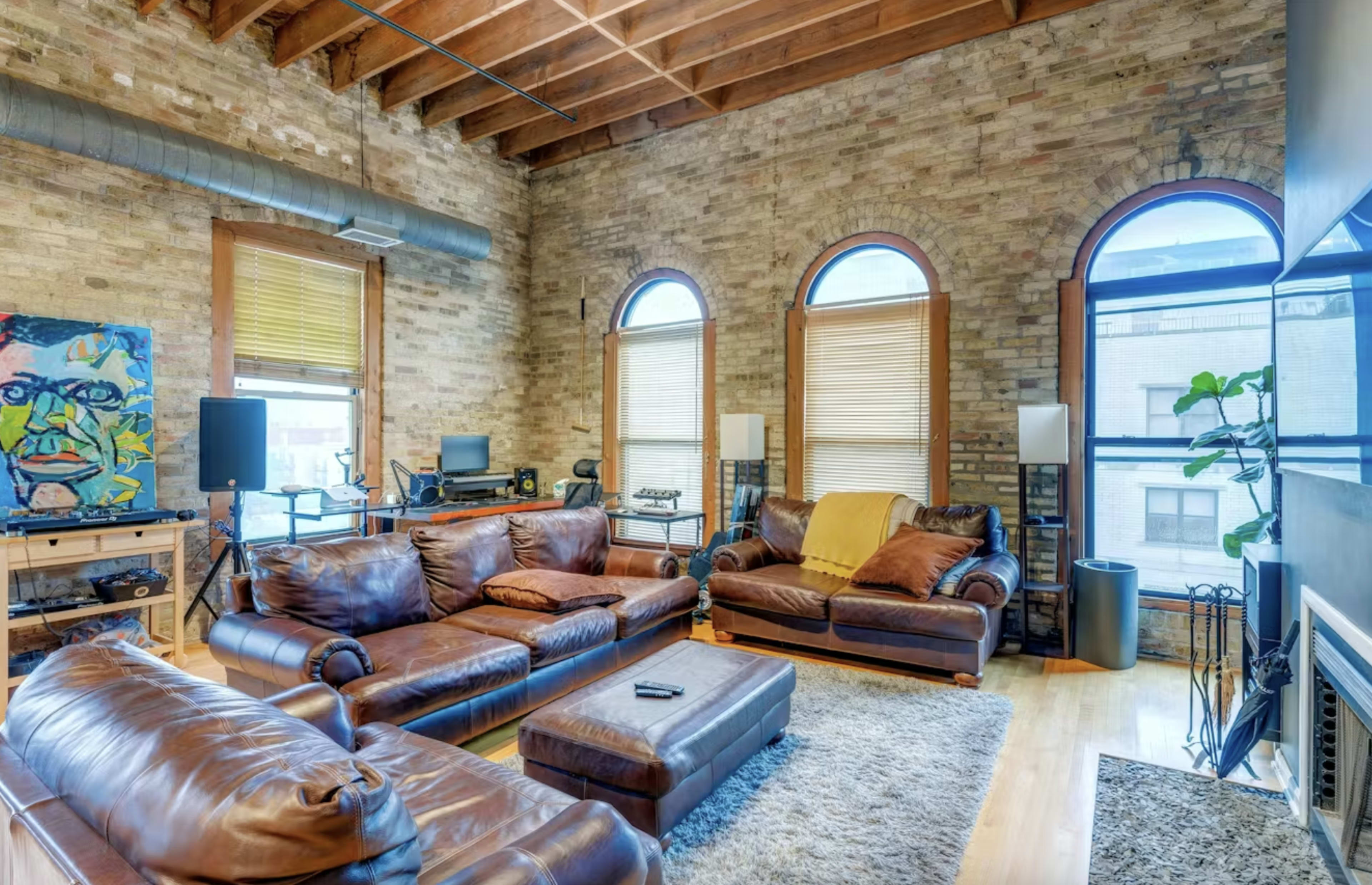 The image shows a spacious living room with exposed brick walls, large arched windows, and leather sofas arranged around a central area rug.