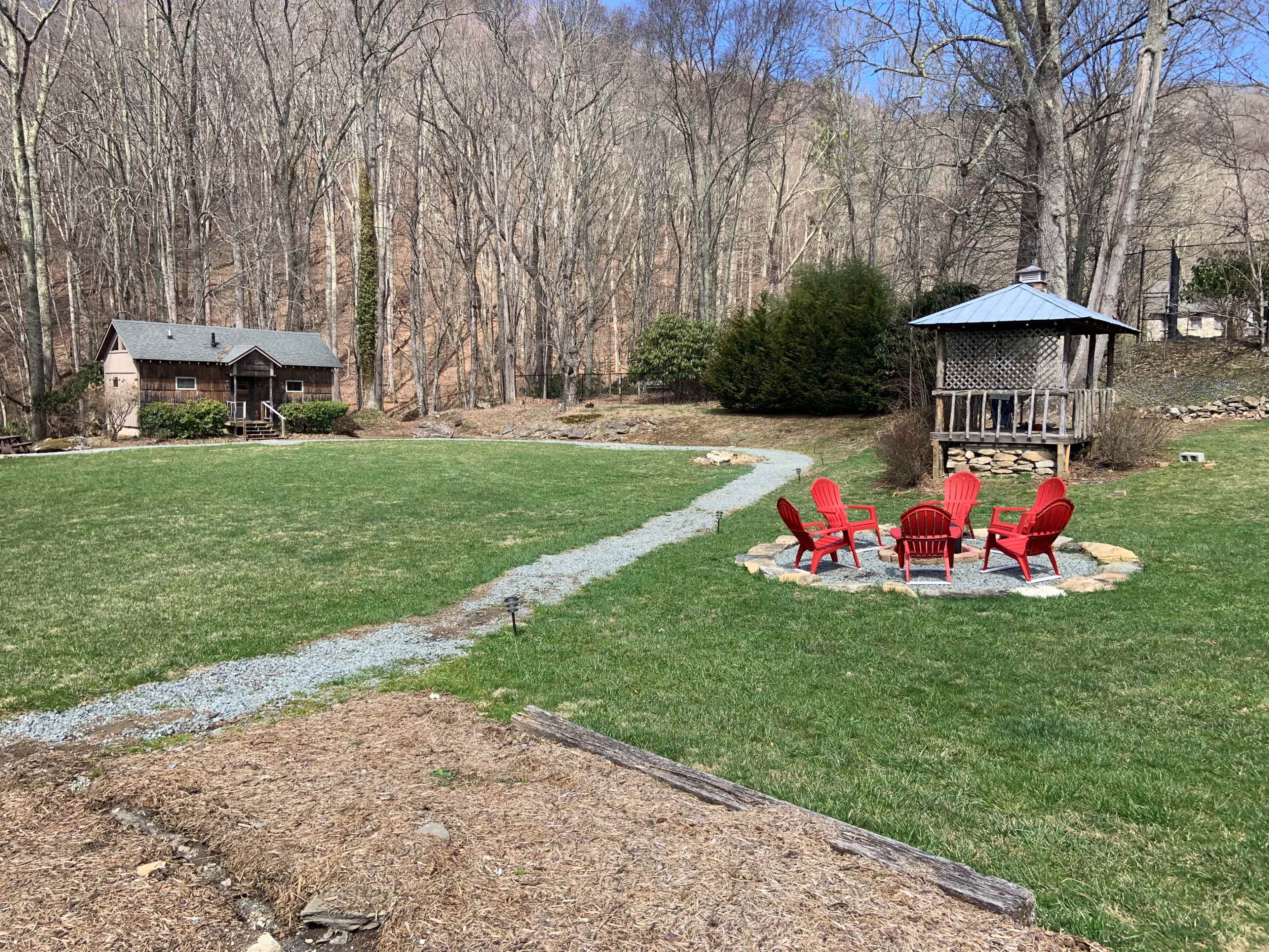 A grassy area with a stone path leading to a circular fire pit surrounded by red chairs, with a cabin in the background and trees beyond.