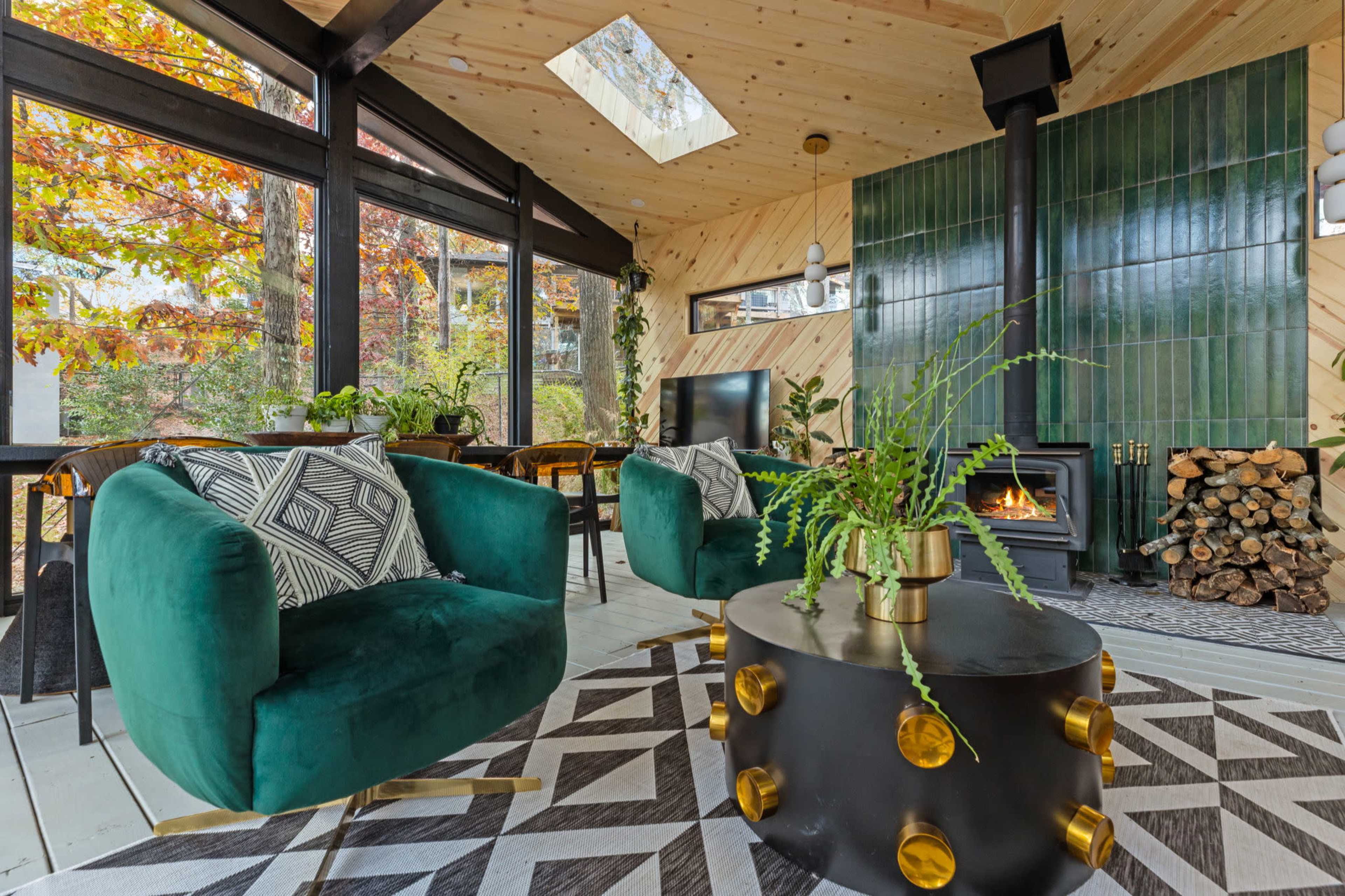 The image showcases a modern living room with two green armchairs, a round coffee table, a wood-burning stove, and large windows letting in natural light, surrounded by autumn foliage.