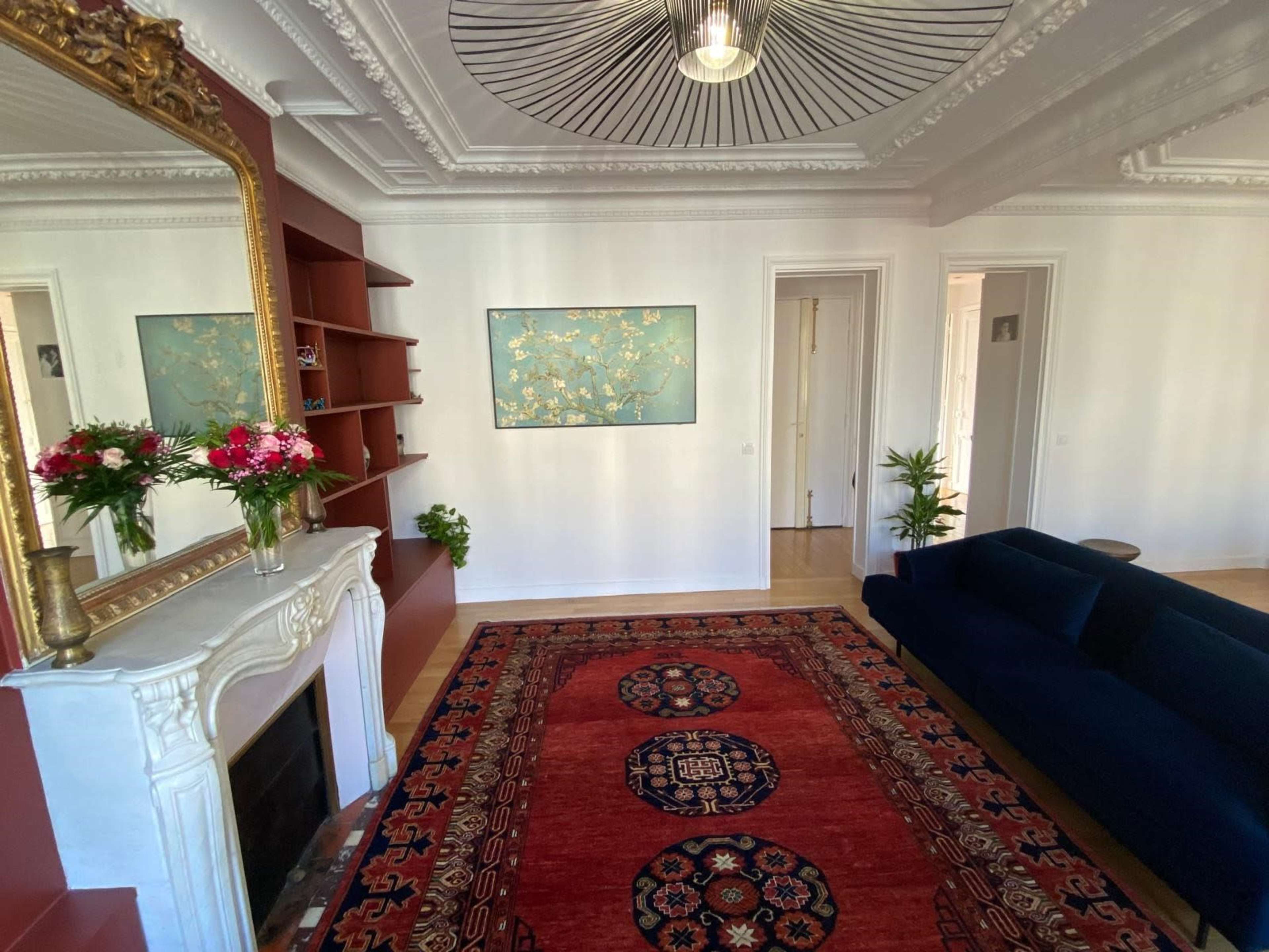 The image shows a stylish living room with a blue sofa, a decorative rug, a large mirror, and a wall adorned with a painting and shelves.